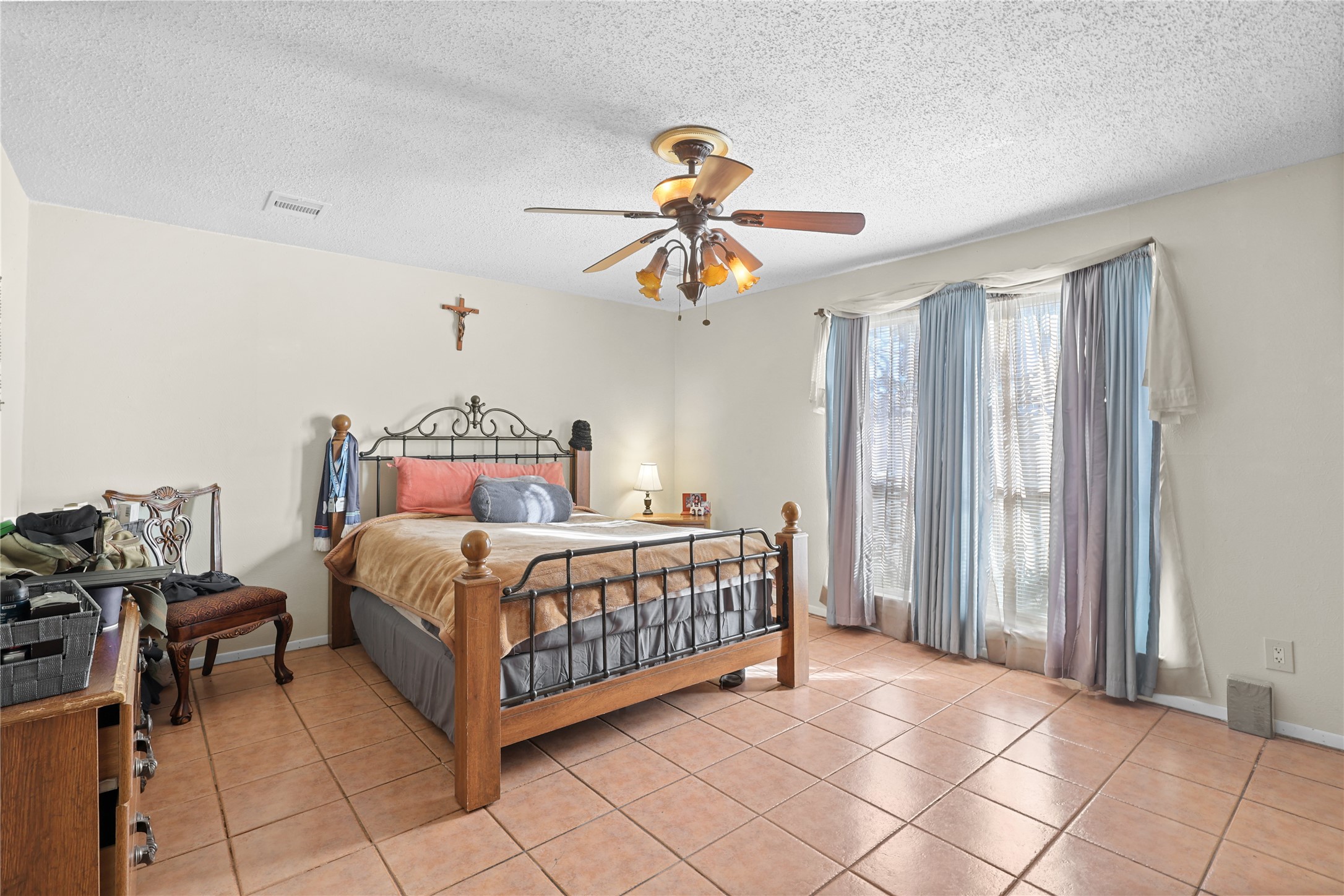 4123 Yupon Ridge Drive Houston, TX 77072 - Photo 9 of 22 This cozy bedroom features a wrought iron bed, tiled flooring, and a ceiling fan. Natural light streams in through large windows with blue curtains, creating a warm and inviting atmosphere.