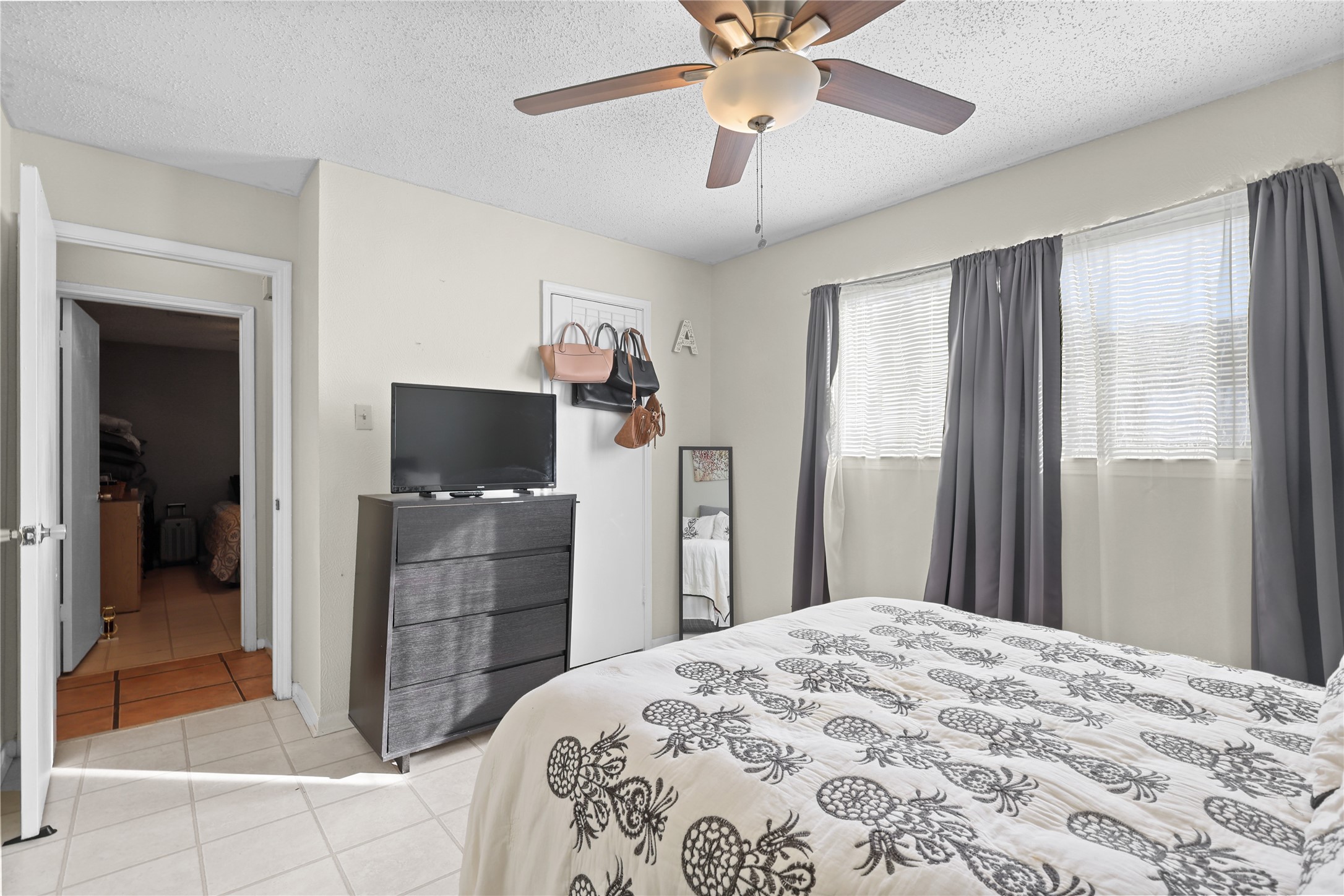 4123 Yupon Ridge Drive Houston, TX 77072 - Photo 10 of 22 Cozy bedroom with natural light from two windows, featuring neutral walls, a ceiling fan, and tiled flooring. Includes a TV on a dark dresser, a full-length mirror, and an adjacent open door leading to another room.
