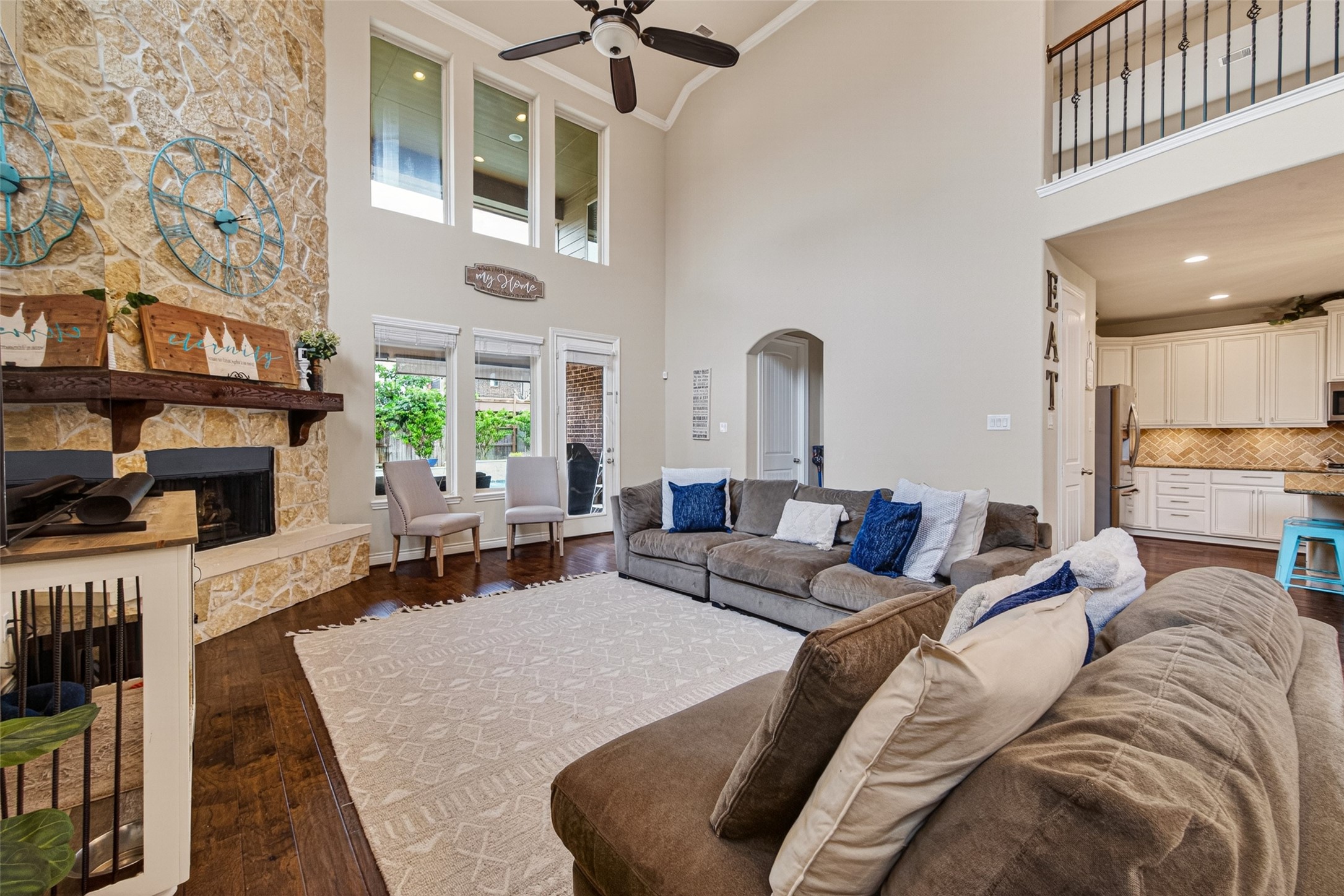 25611 Ellis Ridge Court Katy, TX 77494 - Photo 8 of 49 This spacious living room features a high ceiling with a ceiling fan and an elegant stone fireplace. It has an open concept connecting to a bright kitchen with white cabinetry. Large windows and a cozy sectional offer a welcoming atmosphere, while the hardwood floors and contemporary decor add warmth and style.