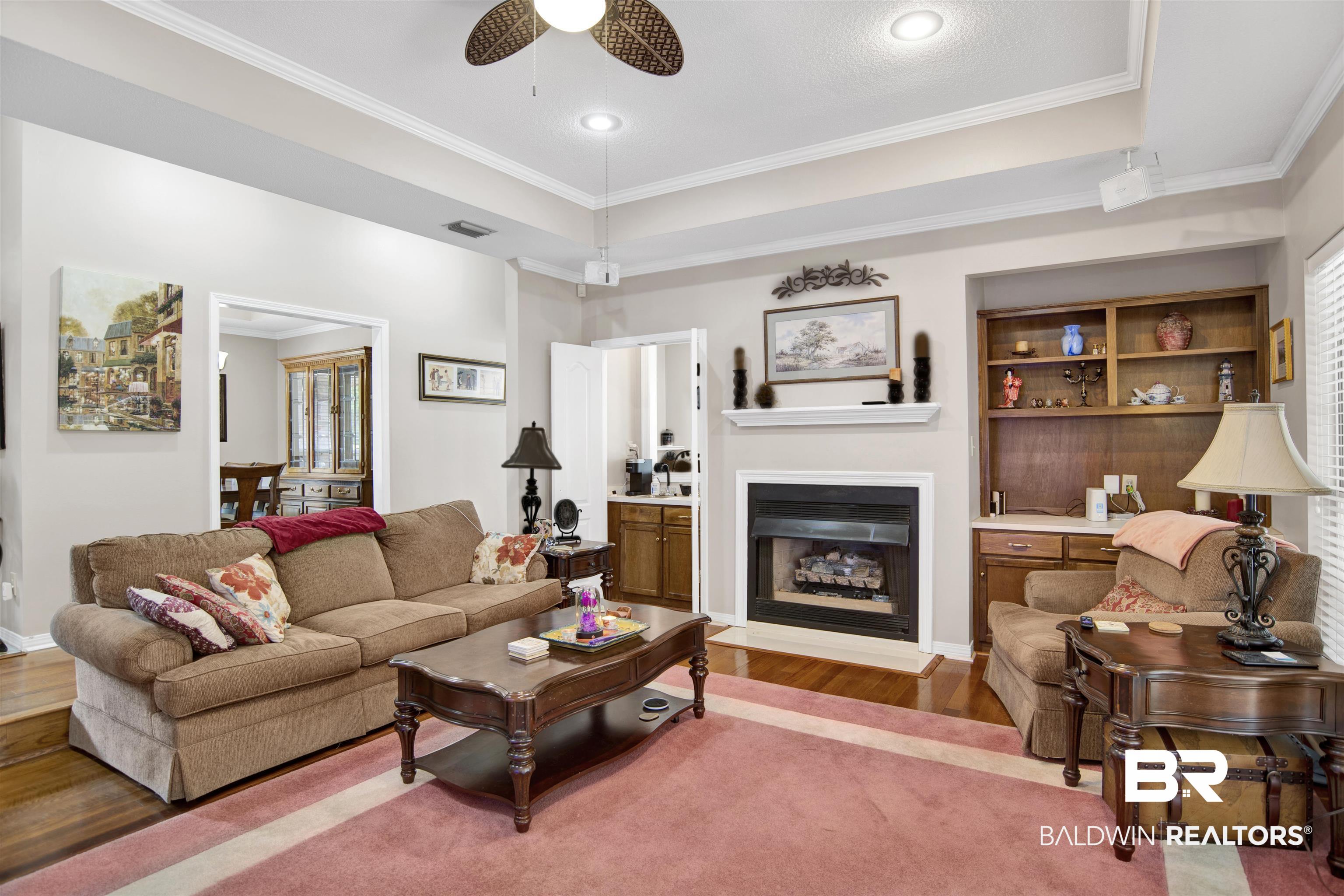 107 Virginia Circle Daphne, AL 36526 - Photo 10 of 47 Living area featuring a fireplace with a mantel, built-in shelving, wood-finish flooring, and a tray ceiling with crown molding