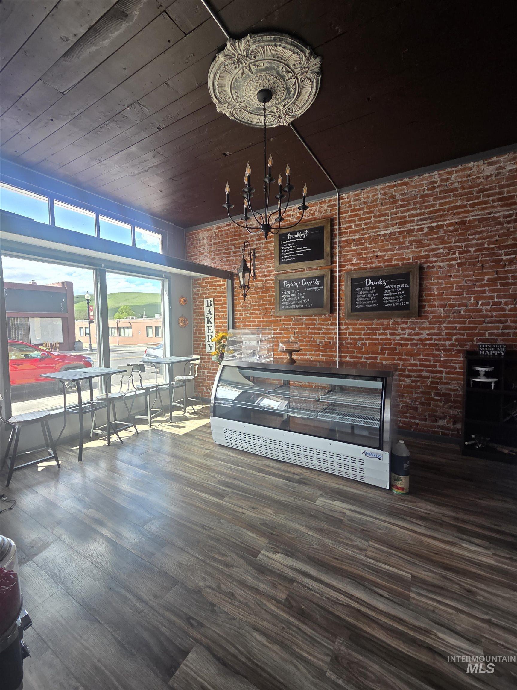 Spacious commercial interior featuring exposed brick walls, wood-finish flooring, and a dark-stained wood plank ceiling with a decorative ceiling medallion and chandelier