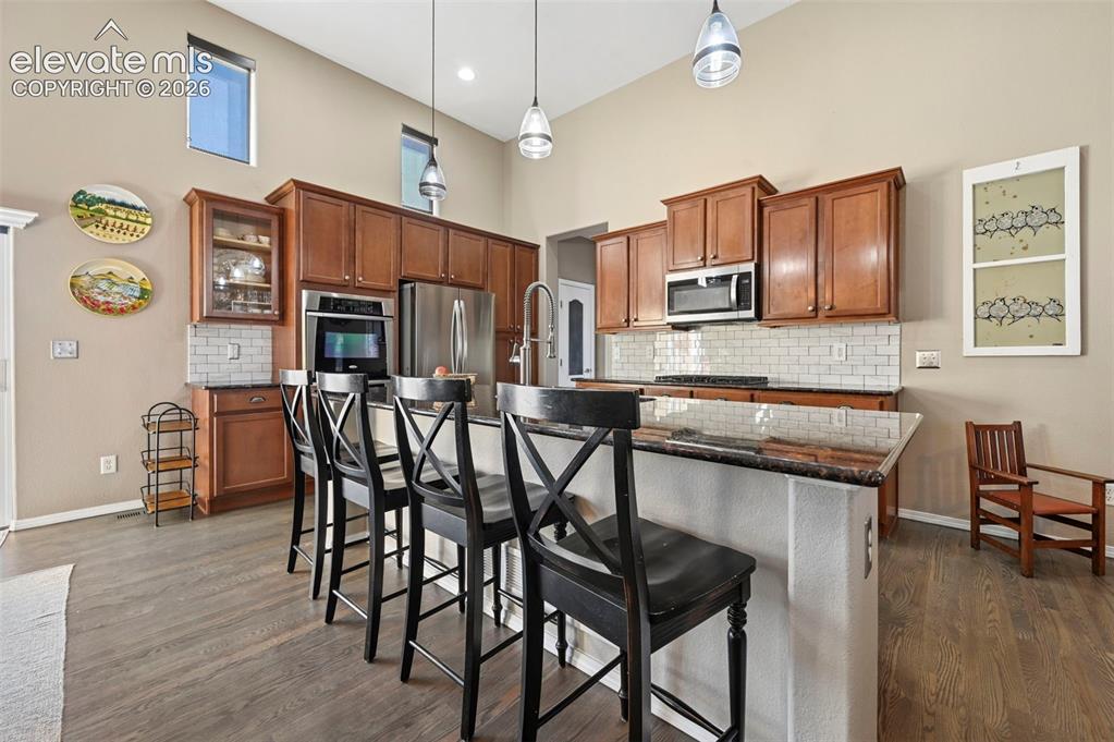 7216 Sylamore Way Peyton, CO 80831 - Photo 6 of 30 Contemporary kitchen featuring wood-finish flooring, warm-toned cabinetry, stainless steel appliances, a subway tile backsplash, and a large central island with a multi-level countertop