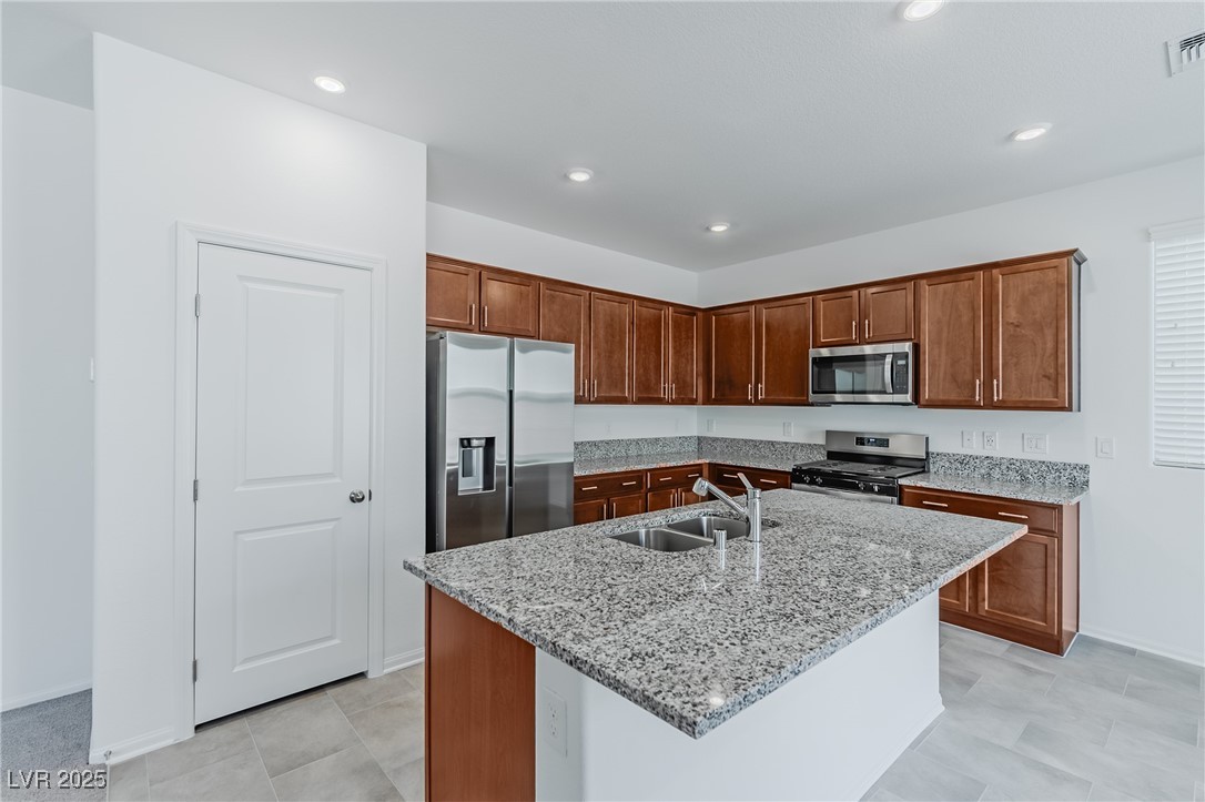 7859 Lacemaker Court Las Vegas, NV 89113 - Photo 3 of 24 Kitchen featuring light stone counters, stainless steel appliances, a kitchen island with sink, recessed lighting, and brown cabinetry
