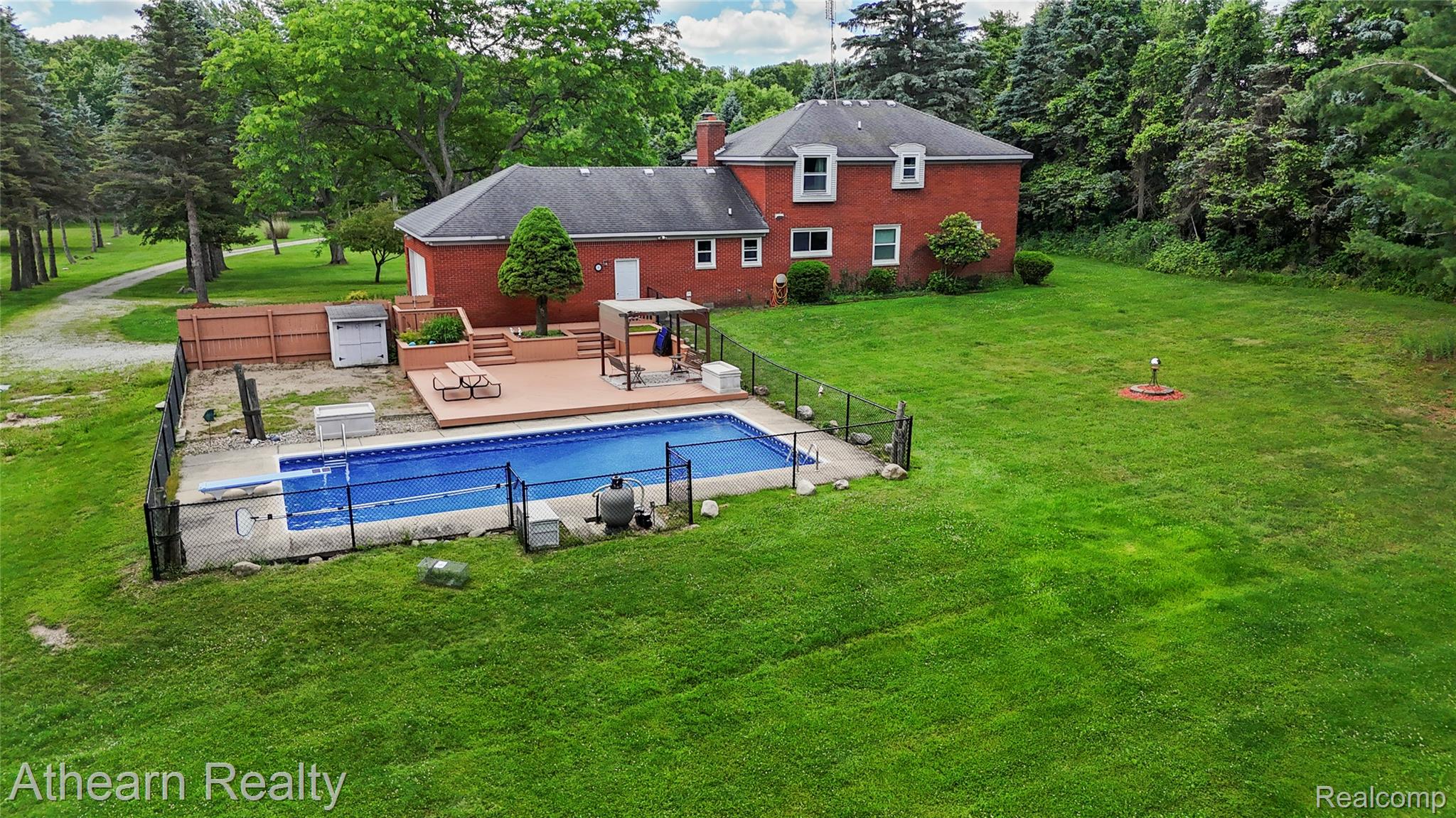 12427 Foley Road Fenton, MI 48430 - Photo 8 of 47 Red brick exterior with white trim, featuring a private fenced swimming pool, extensive green lawn, tiered wood deck, and a gravel driveway