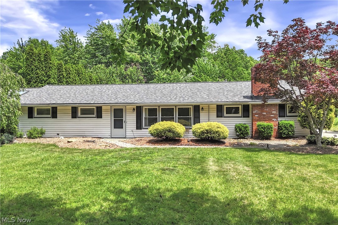 Ranch-style house featuring light-colored siding, a gray shingle roof, dark window shutters, a brick chimney, and mature landscaping.