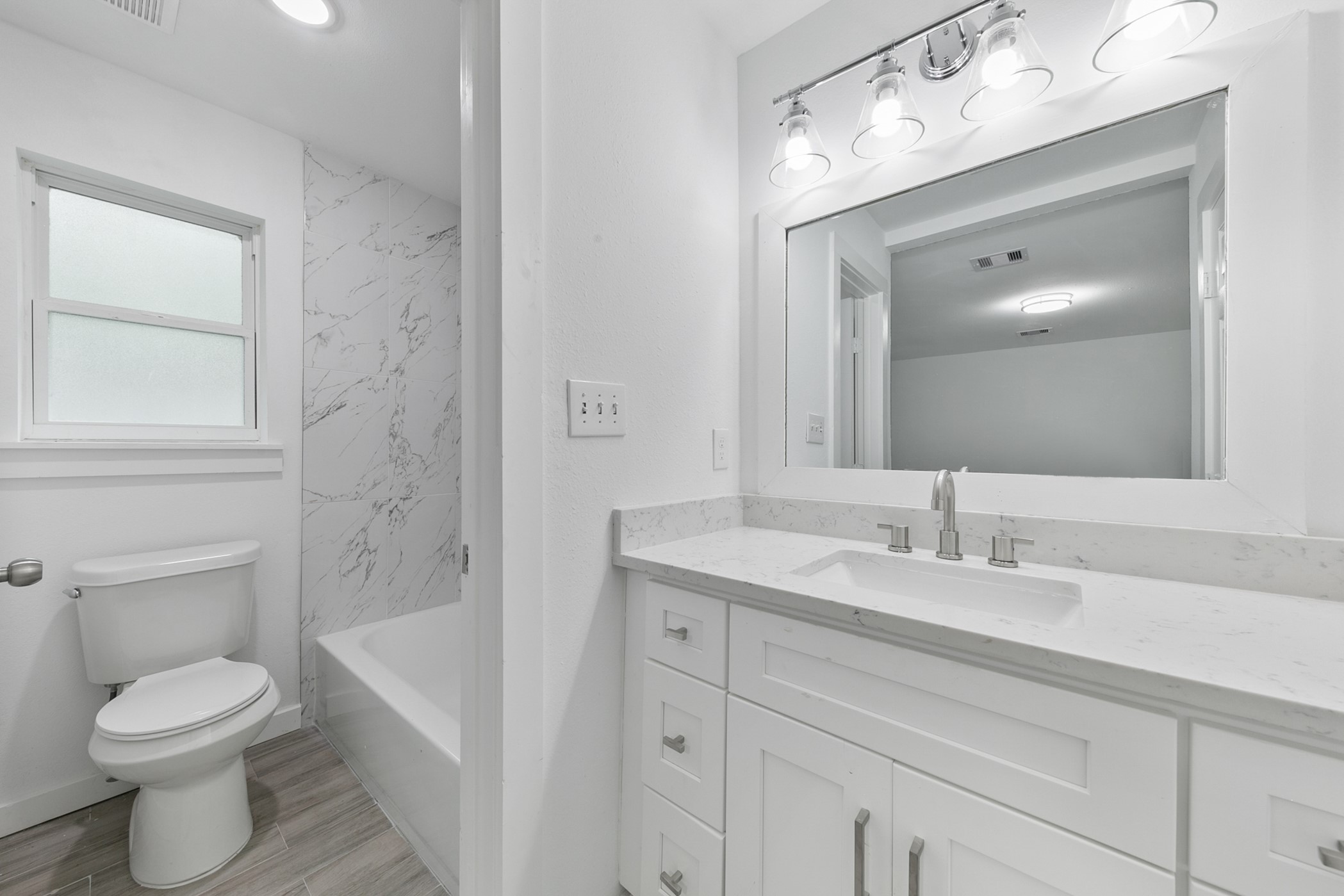 6811 McCullum Road Houston, TX 77489 - Photo 12 of 22 This modern bathroom features sleek white cabinetry with a spacious countertop, a large mirror with bright lighting, a bathtub with elegant marble-patterned tiles, and a window for natural light.
