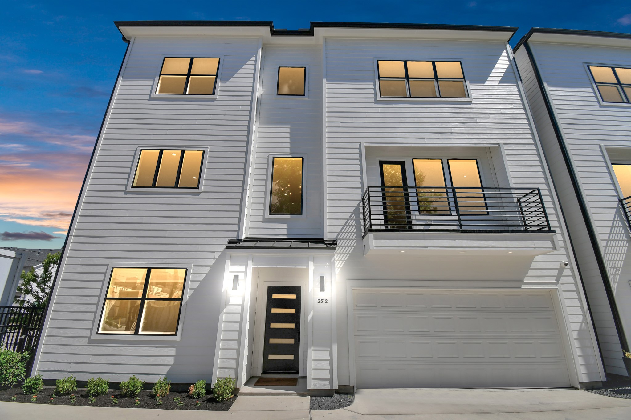 This modern, three-story townhouse features sleek white siding, large windows for ample natural light, and a balcony. It includes an attached two-car garage and a stylish front entrance, perfect for contemporary living.