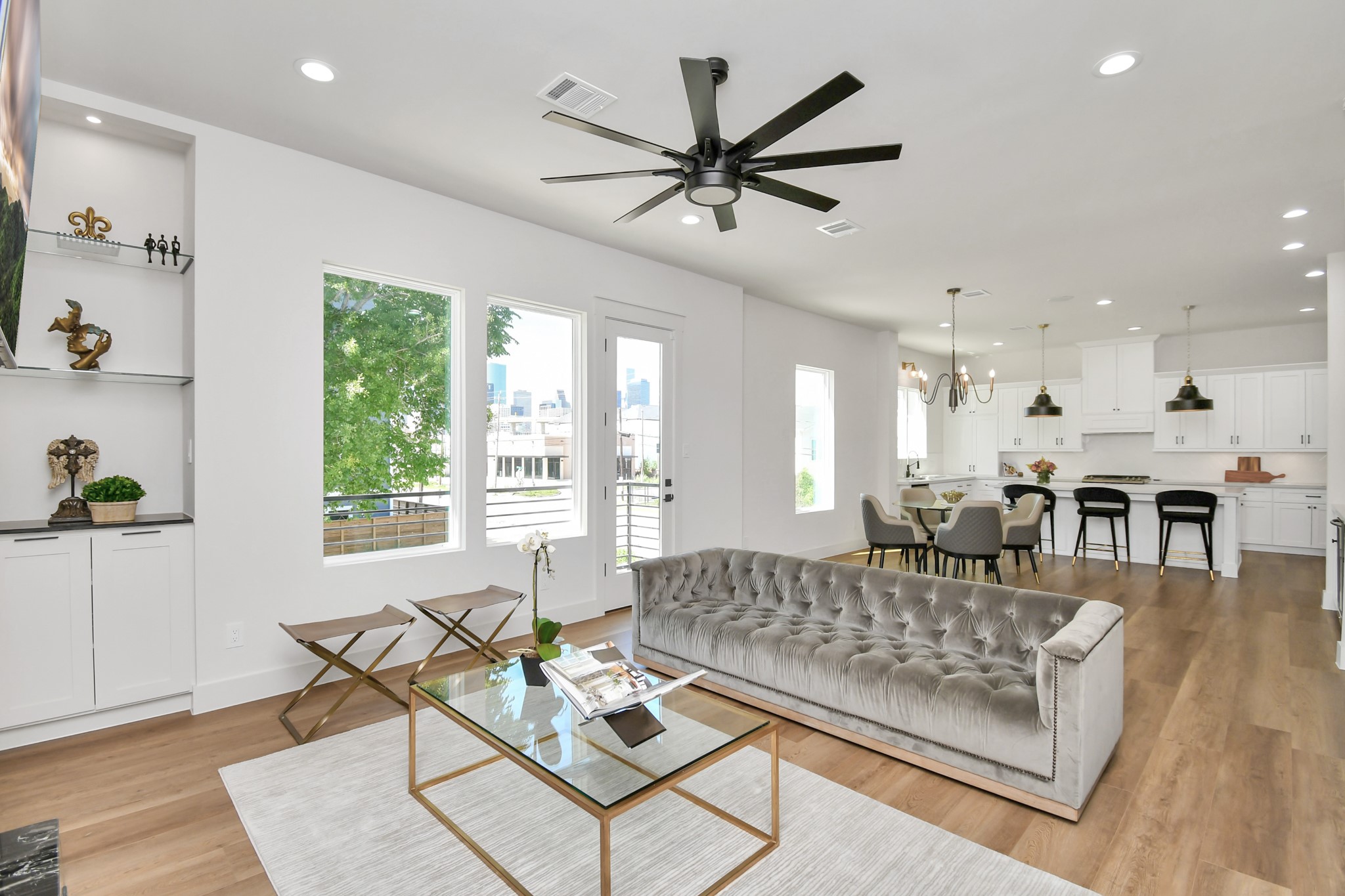 2512 Gray Street Houston, TX 77003 - Photo 13 of 46 This photo shows a bright, open-concept living and dining area with modern finishes. It features a plush gray sofa, a glass coffee table, and sleek shelves. The kitchen has white cabinetry, a large island with bar seating, and stylish pendant lighting. Large windows offer plenty of natural light and a view of the outside.
