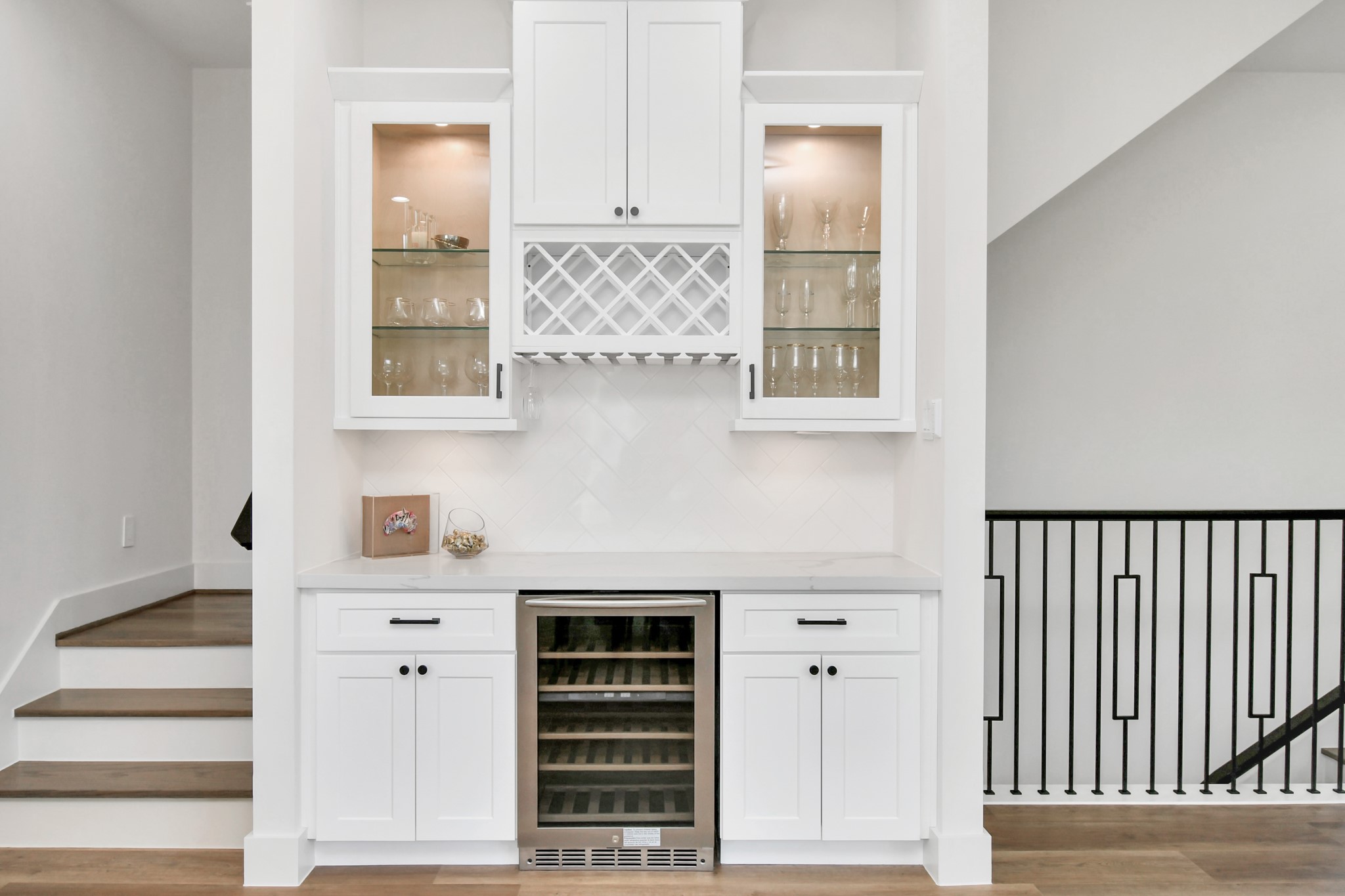 2512 Gray Street Houston, TX 77003 - Photo 17 of 46 This photo showcases a stylish built-in bar area with white cabinetry, glass display shelves, and a wine rack. The space includes a wine cooler and is accented with subtle lighting. It's conveniently situated near a staircase and features a modern railing design.