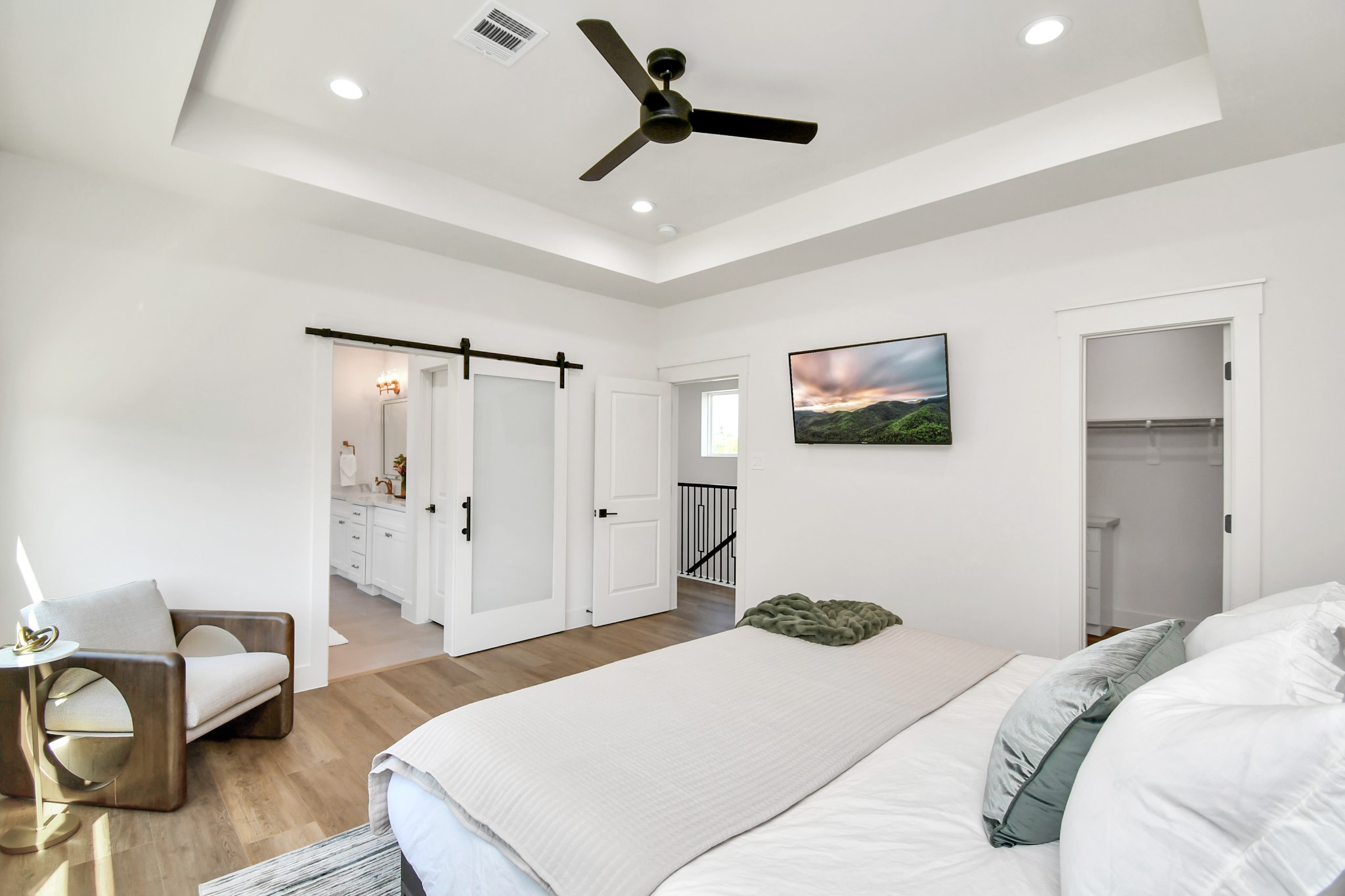2512 Gray Street Houston, TX 77003 - Photo 26 of 46 This is a modern, bright bedroom featuring a tray ceiling, recessed lighting, and a ceiling fan. It includes a comfortable bed, a cozy chair, and a mounted TV. The room has a sliding barn door leading to an en-suite bathroom and a walk-in closet, with wood flooring throughout.