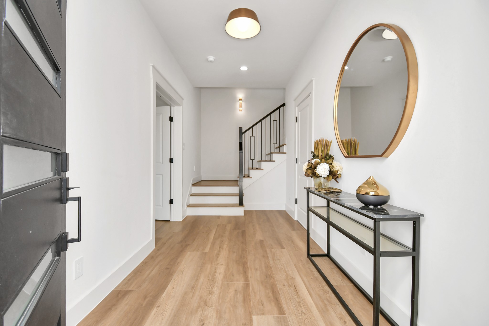 2512 Gray Street Houston, TX 77003 - Photo 3 of 46 This photo shows a modern, welcoming entryway with light wood flooring and white walls. It features a sleek black front door, a stylish console table with decorative items, and a large round mirror. A staircase with a contemporary railing leads to the upper level, creating a spacious and elegant atmosphere.