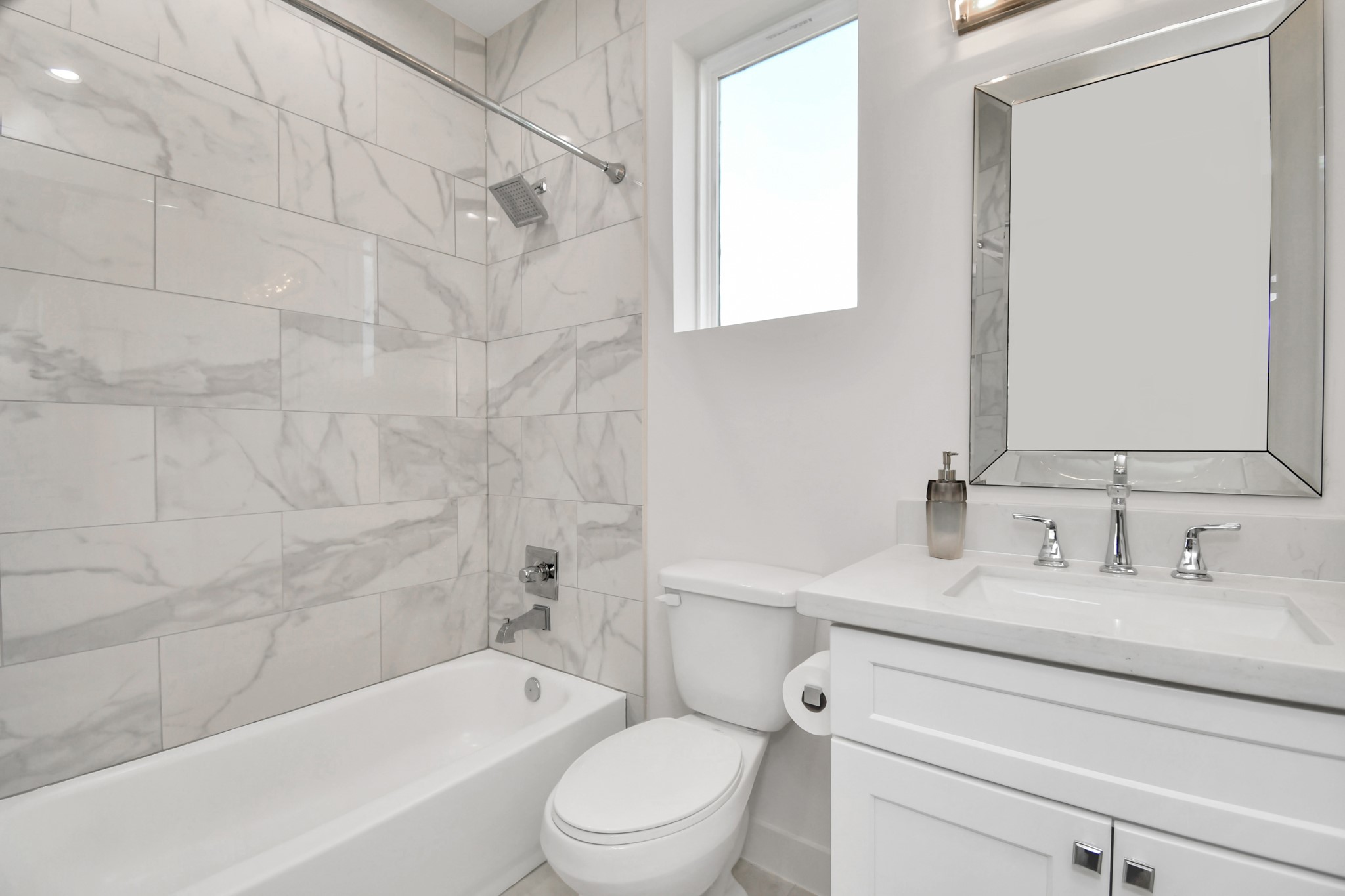 2512 Gray Street Houston, TX 77003 - Photo 6 of 46 This bathroom features a modern design with sleek marble tile walls, a bathtub with a shower, a white vanity with a mirrored frame, and a bright window for natural light.