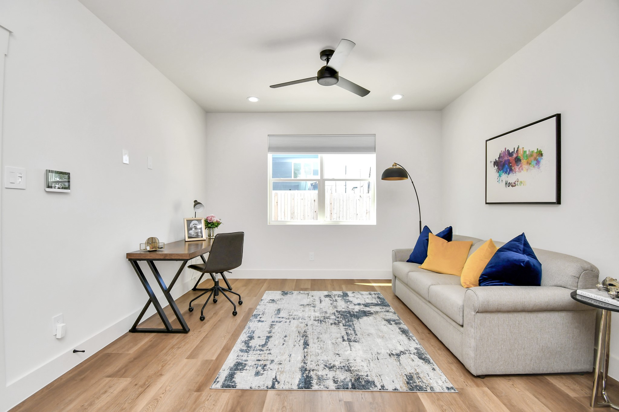 2512 Gray Street Houston, TX 77003 - Photo 7 of 46 This inviting room features a modern, minimalist design with a comfortable sofa accented by colorful pillows, a sleek desk, and a stylish ceiling fan. The space is well-lit with natural light from the window, highlighting the wooden flooring and vibrant wall art. Perfect for a home office or cozy relaxation area.