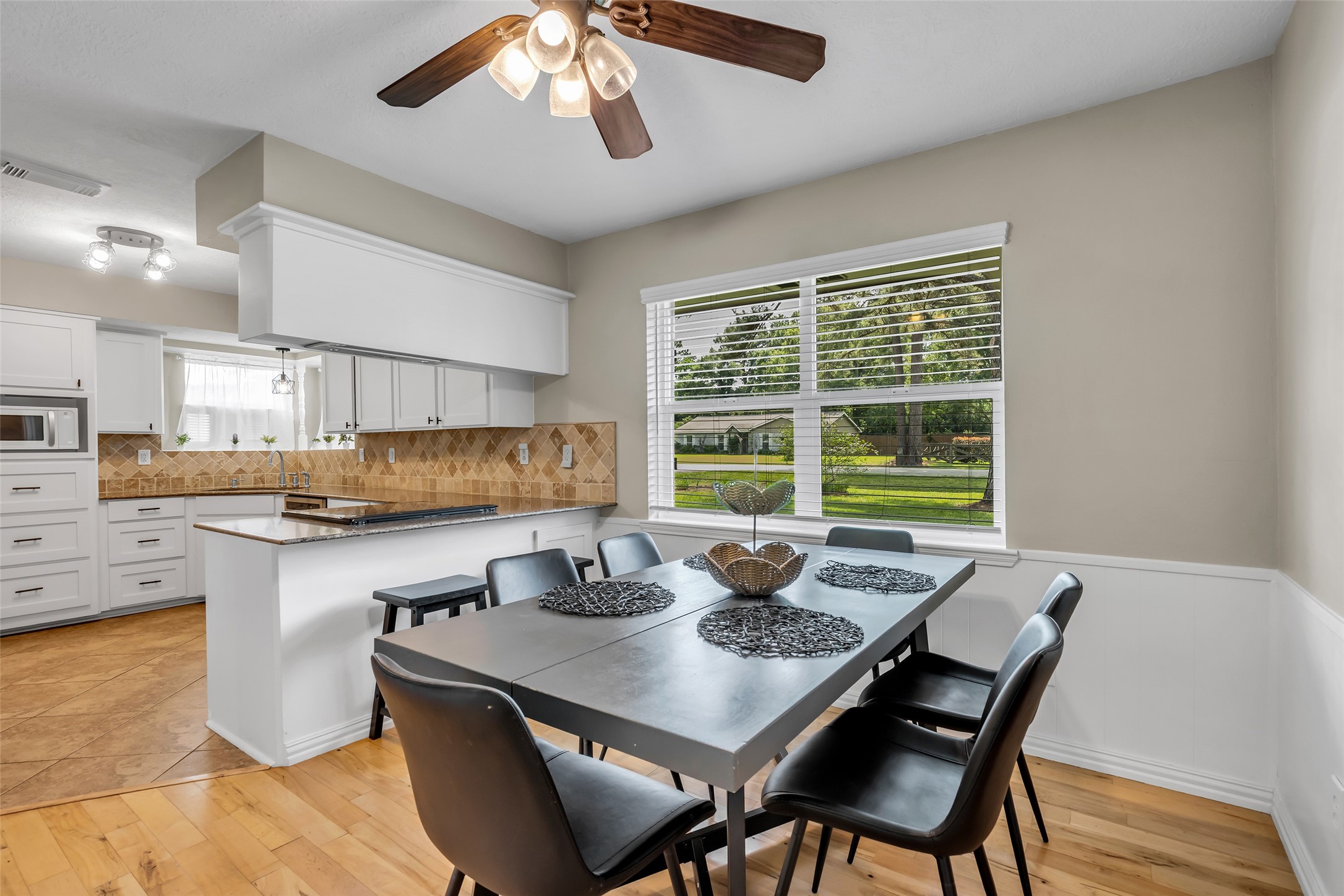 22502 Fields Lane Spring, TX 77389 - Photo 10 of 39 Bright dining area with a modern table and chairs, featuring a large window offering a view of the garden. Adjacent is a kitchen with white cabinetry, tile backsplash, and a ceiling fan for added comfort.