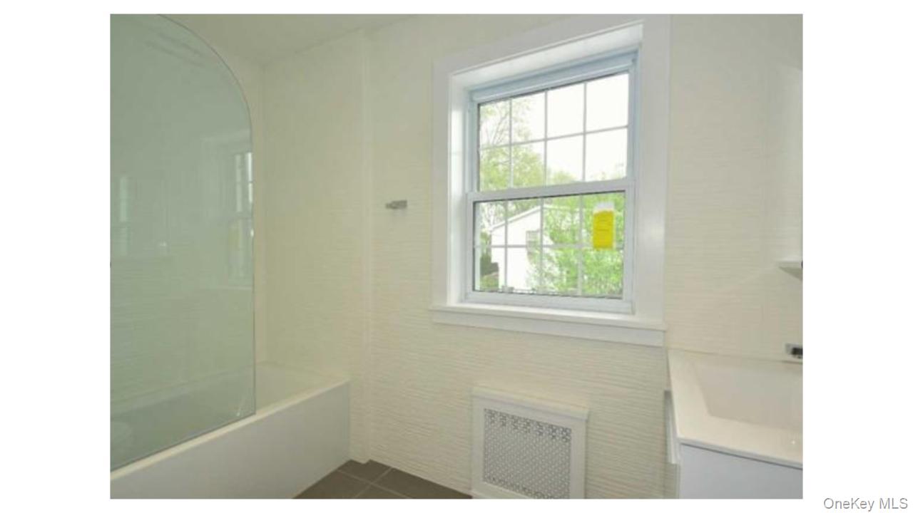 2 Arleigh Road Great Neck, NY 11021 - Photo 7 of 22 White bathtub with a curved glass shower partition, white window with grid panes, textured wall covering, and a white vanity with an integrated sink