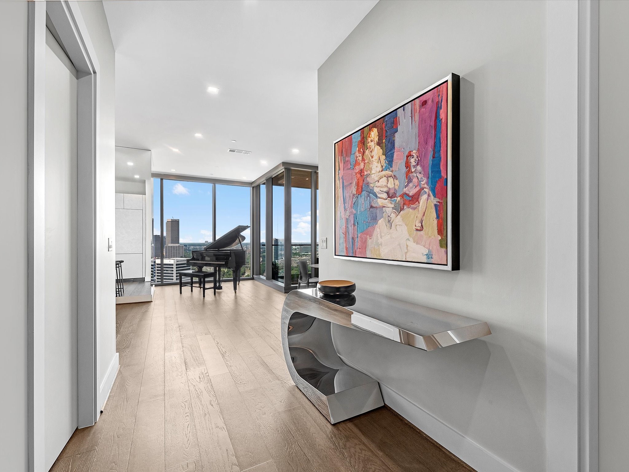 This modern, bright hallway features sleek wood flooring, a contemporary console table, and vibrant artwork. It leads to an open living area with large windows offering stunning city views and natural light.