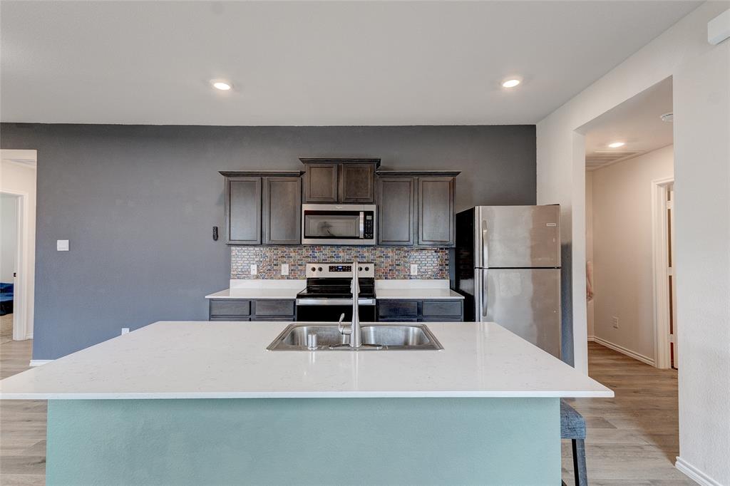 1016 Rio Bravo Drive Forney, TX 75126 - Photo 6 of 25 Kitchen island with a built-in sink and white countertop, dark wood-finish cabinetry, stainless steel appliances, and a colorful tile backsplash