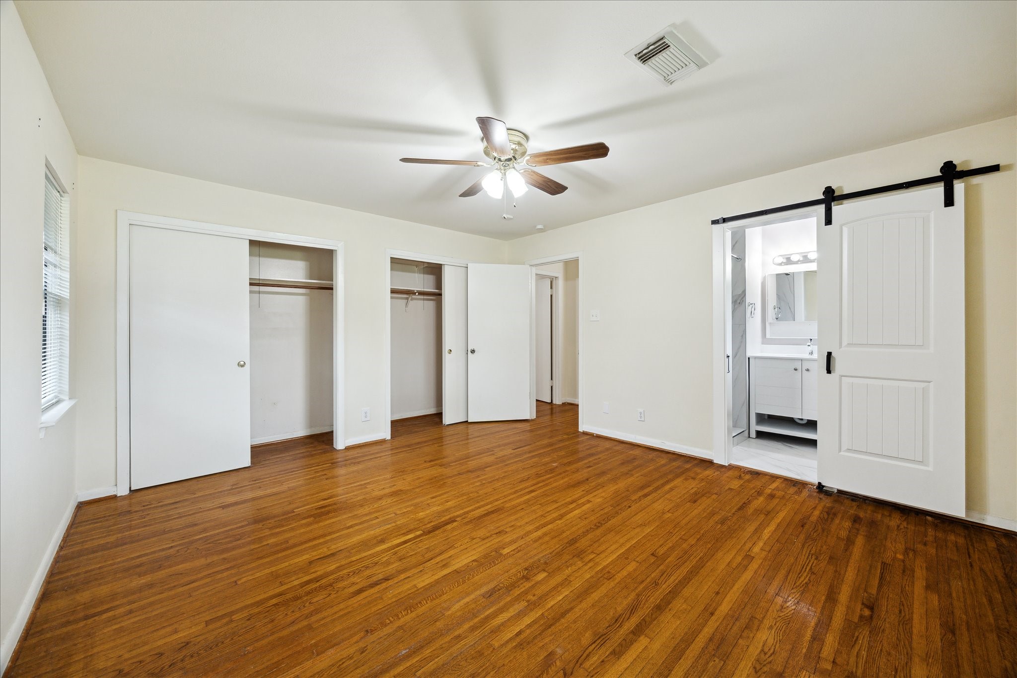 8907 Autauga Street Houston, TX 77080 - Photo 12 of 20 This photo shows a spacious bedroom with hardwood floors, a ceiling fan, and two large closets. There's a modern ensuite bathroom accessible through a stylish sliding barn door. Natural light enters through a window, creating a bright and welcoming space.