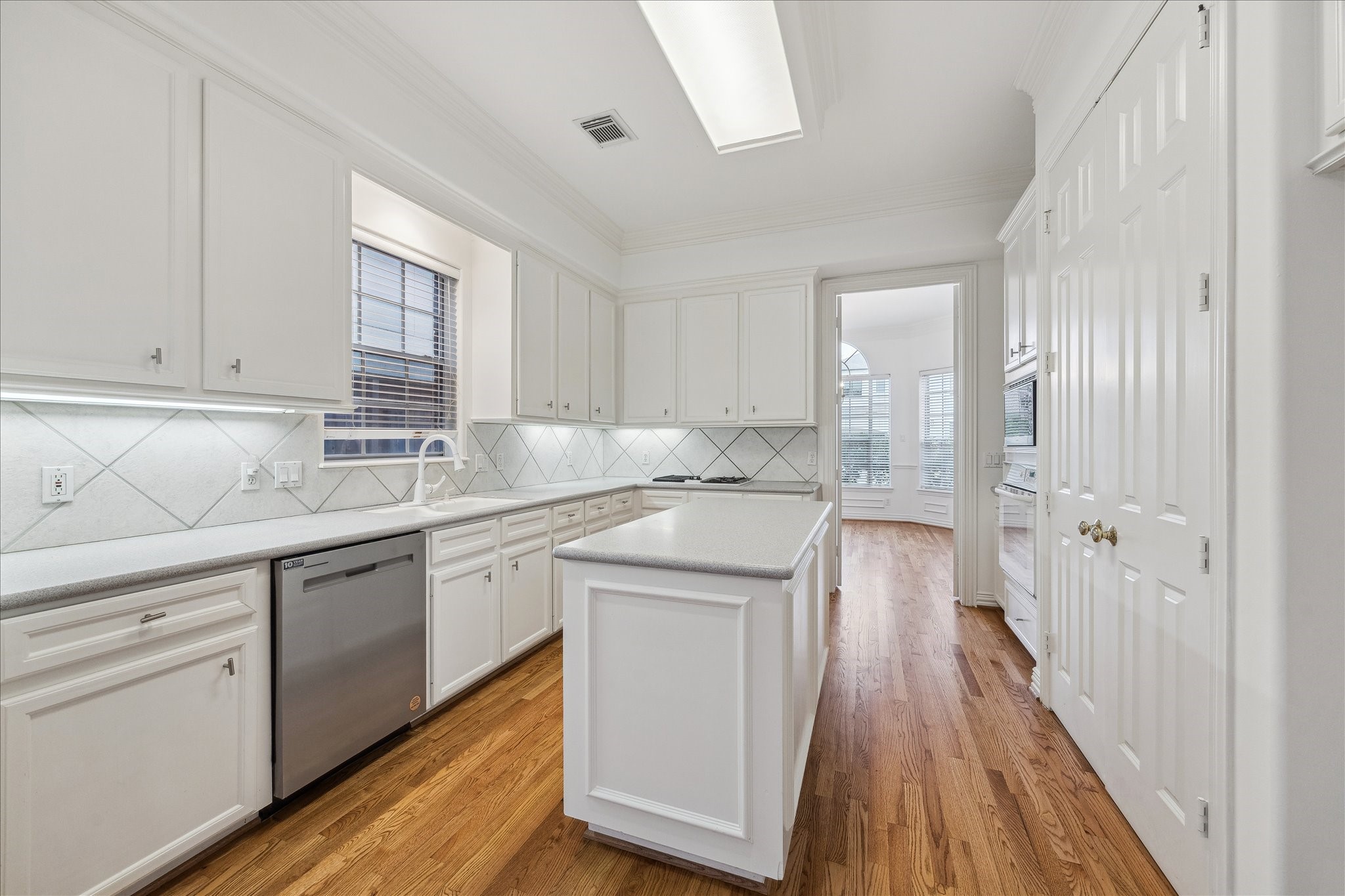 5107 Locust Street Bellaire, TX 77401 - Photo 20 of 45 Light-filled kitchen featuring extensive white cabinetry, crown molding, and a central island designed for prep and serving. Crisp tile backsplash, under-cabinet lighting, and great counter space create a clean, functional workspace. Stainless dishwasher, built-in appliances, and a skylight enhance both efficiency & natural light, with gorgeous hardwood floors.