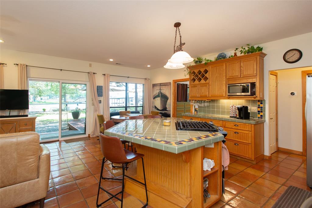 121 Xit Ranch Road Trinidad, TX 75163 - Photo 8 of 40 Kitchen and living area featuring terracotta tile flooring, an island with a tiled countertop and cooktop, wooden cabinetry, and glass sliding doors leading to the back covered deck and lake area.
