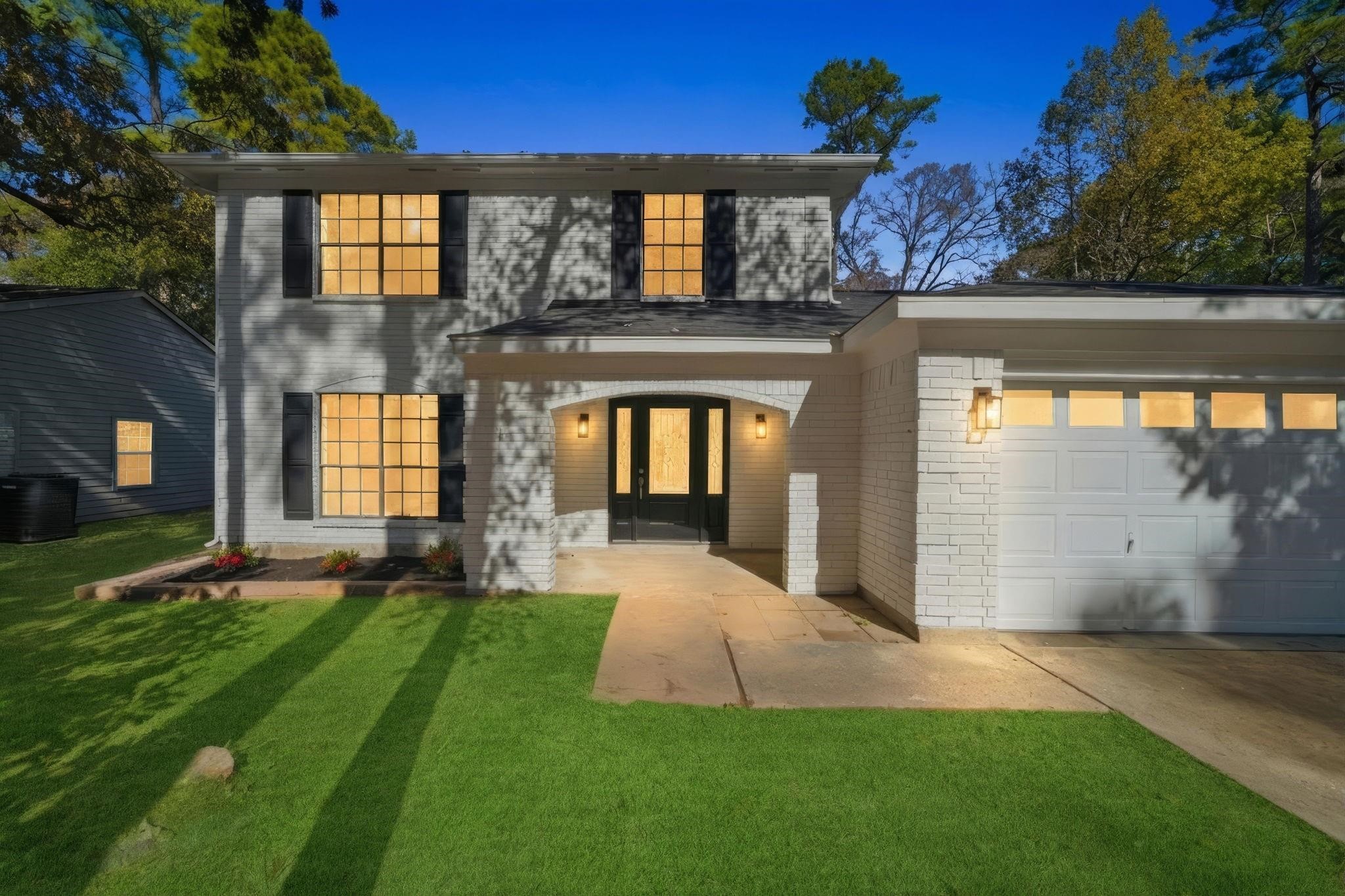 This photo shows a charming two-story brick home with a modern look. It features a well-maintained front yard, a welcoming entrance with double doors, large windows, and an attached garage. The exterior lighting adds warmth and appeal.