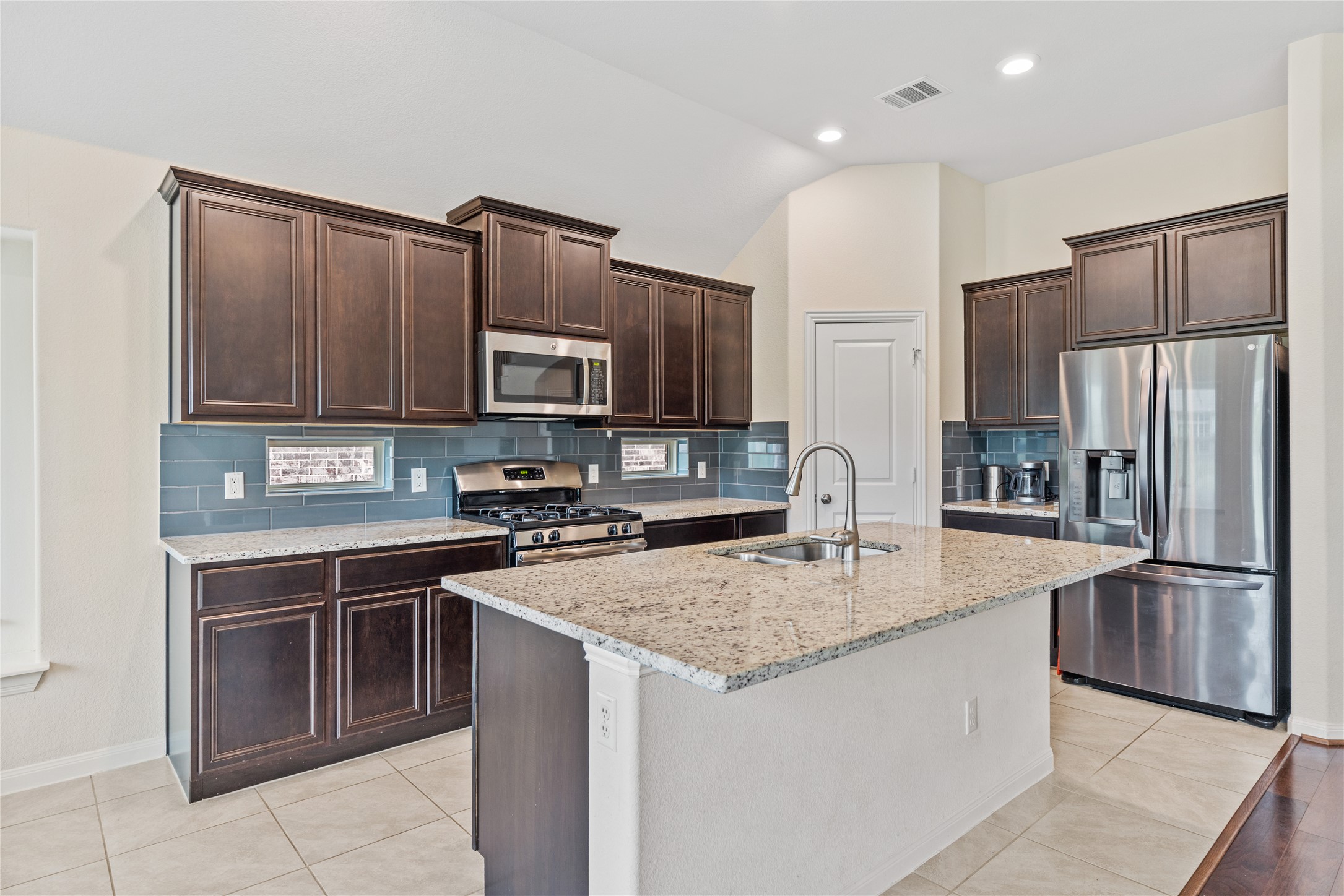 5012 Yucca Bloom Drive Georgetown, TX 78626 - Photo 6 of 35 Kitchen featuring an island with an undermount sink and gooseneck faucet, dark wood cabinetry, light-toned tile flooring, stainless steel appliances, and a blue subway tile backsplash