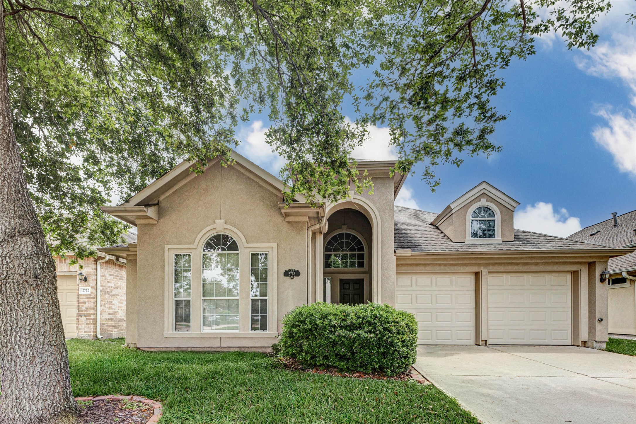 Classic front exterior framed by mature shade trees, manicured lawn and a bright blue sky. The soft stucco façade features arched architectural windows, an elevated front entry, neatly trimmed shrubs and a two-car attached garage. A wide driveway offers convenient parking and approach, while the large tree canopy adds shade and character. The home presents a warm, welcoming first impression with timeless curb appeal, clean lines and distinctive architectural detailing.