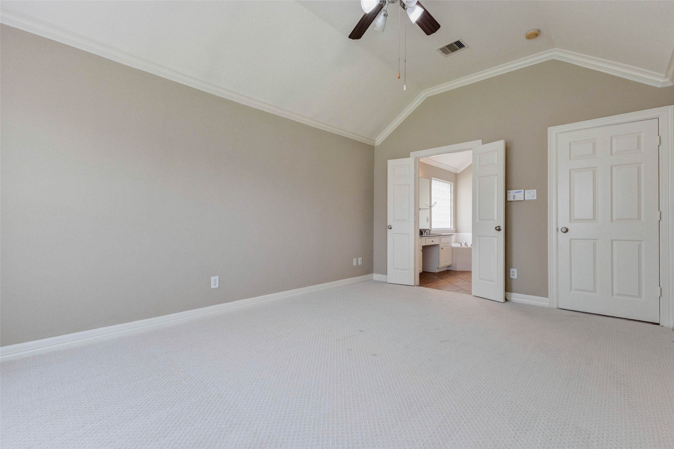 2718 Ridgeglen Lane Houston, TX 77082 - Photo 18 of 26 Primary bedroom view showing a spacious layout, vaulted ceiling, ceiling fan and direct access to the private bath. Double doors open to the en-suite area, where the vanity and soaking tub are visible, while a separate closet door sits nearby. Neutral walls, soft carpet, crown molding and ample floor space create a comfortable retreat with practical convenience and a calm, open feel.