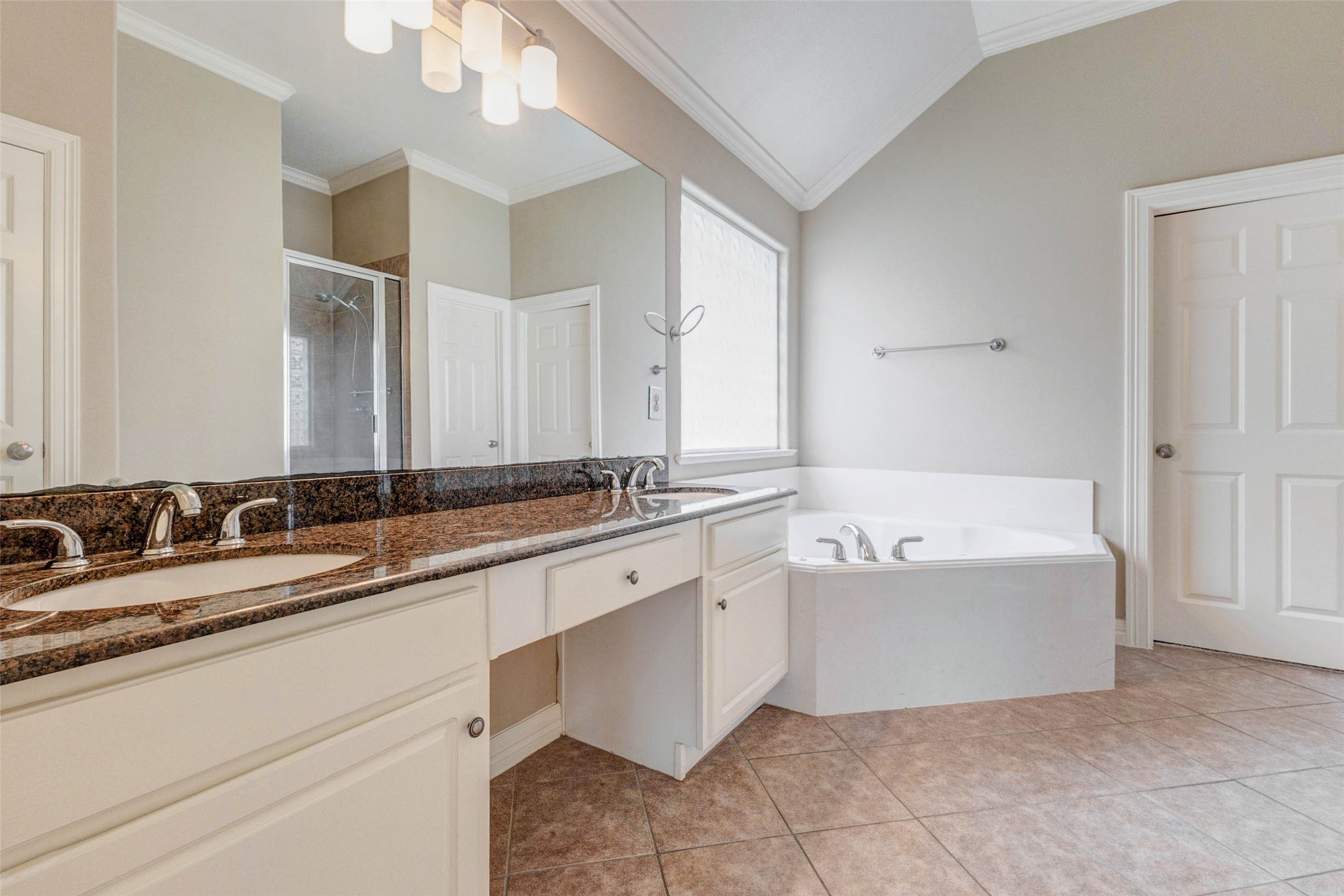 2718 Ridgeglen Lane Houston, TX 77082 - Photo 19 of 26 Spacious primary bath with an extended granite-topped double vanity, framed by a full-width mirror and modern multi-light fixture. The room features tile flooring, neutral walls, vaulted ceiling lines and crisp white trim. A large soaking tub sits beneath a privacy glass-block window, creating a bright, serene focal point, while the separate shower is visible in the mirror reflection. Generous counter space and dual sinks add comfort and everyday convenience.