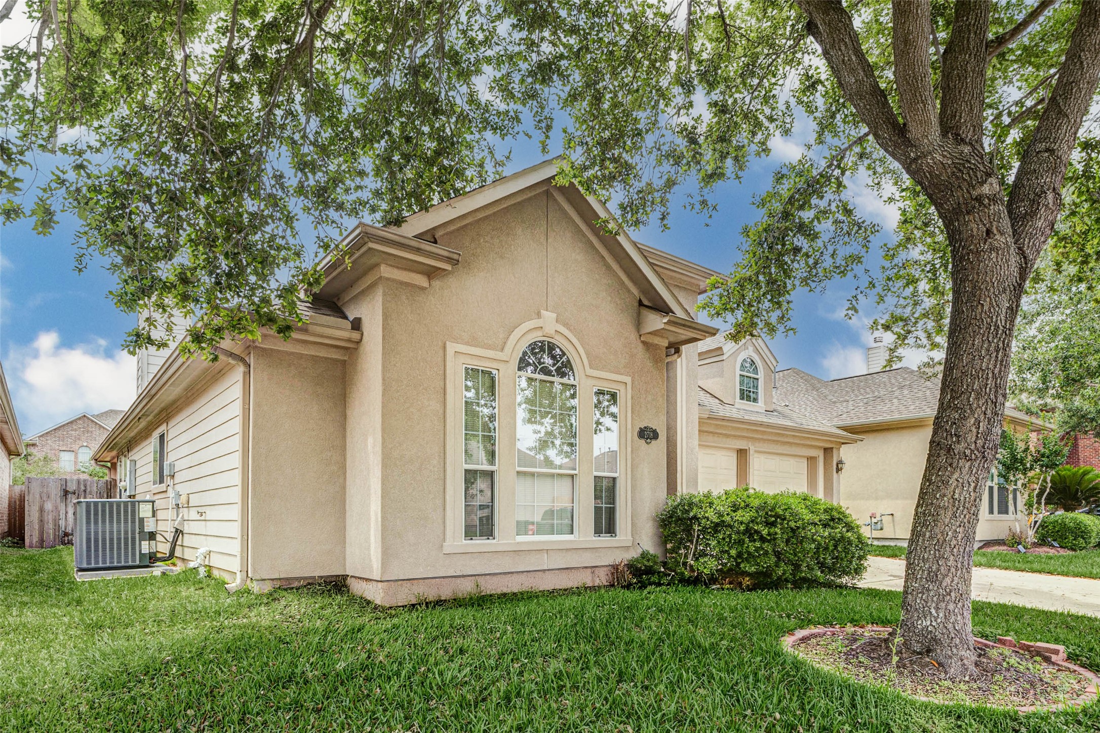 2718 Ridgeglen Lane Houston, TX 77082 - Photo 2 of 26 Side-angle exterior view showcasing the home’s stucco elevation, tall arched front window and mature tree canopy that adds shade and character to the front yard. The lawn wraps around the side of the home, with visible rear fencing and utility equipment along the side yard. This perspective highlights the clean roofline, architectural window trim, tidy landscaping and the single-story profile that makes the exterior feel approachable, attractive and well maintained.
