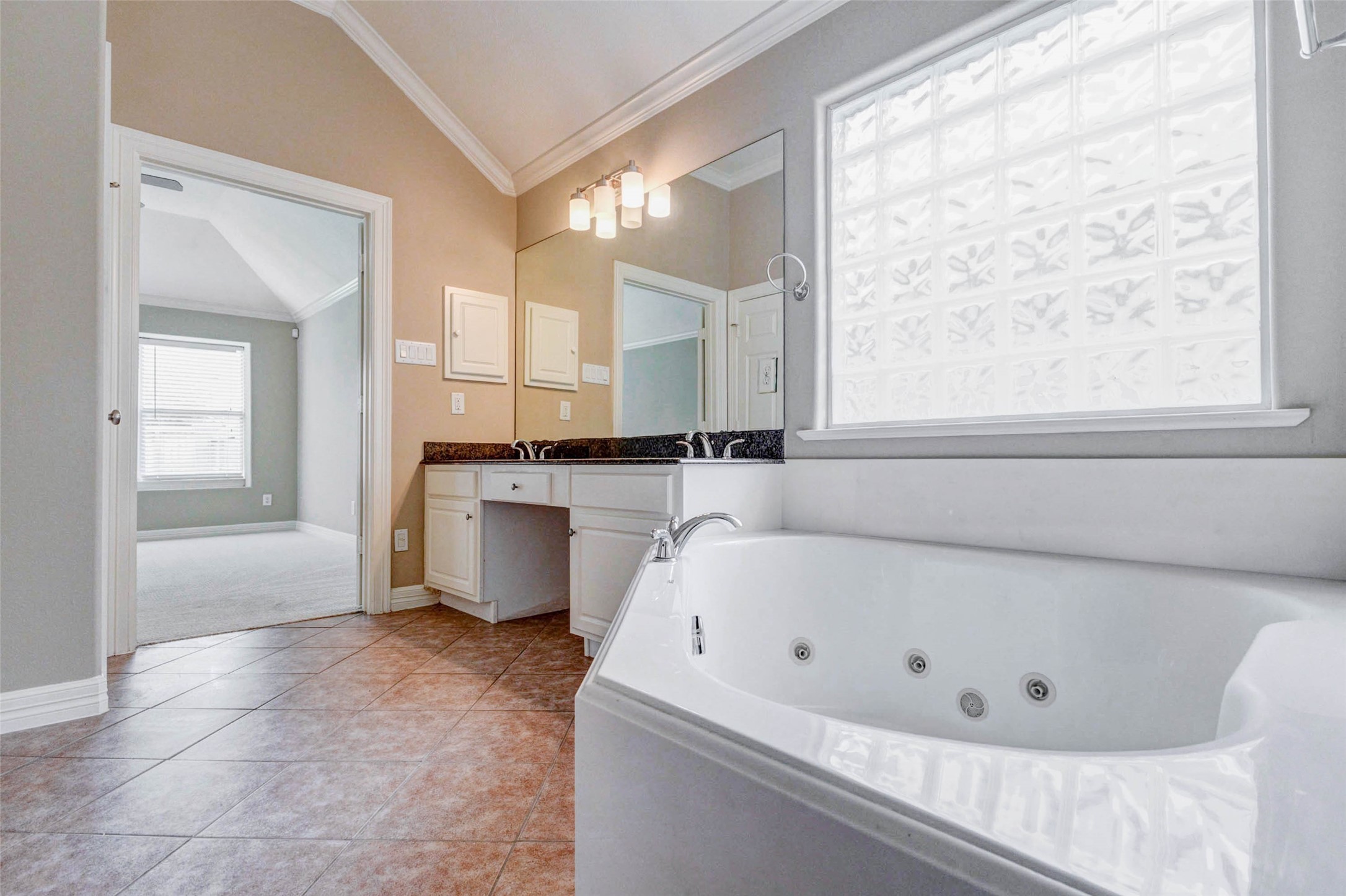 2718 Ridgeglen Lane Houston, TX 77082 - Photo 20 of 26 Primary bathroom view centered on the large soaking tub beneath a glass-block privacy window that filters in natural light. The tile floor, crown molding and neutral finishes create a clean, calming feel, while the long vanity with granite counter and dual sinks adds practical storage and prep space. The open doorway connects directly to the primary bedroom, giving the suite an easy, private flow between sleeping and bath areas.