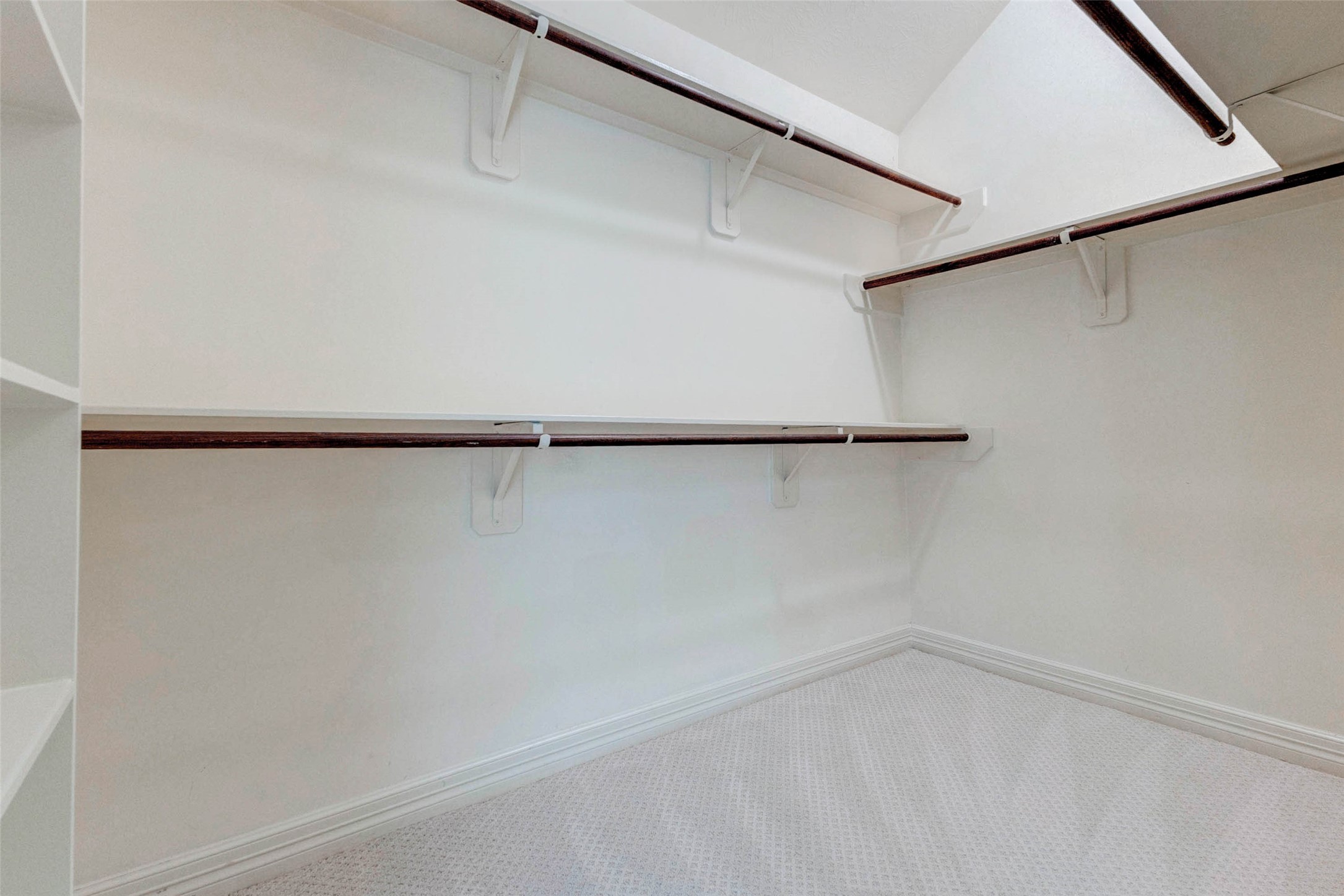 2718 Ridgeglen Lane Houston, TX 77082 - Photo 21 of 26 Walk-in closet with generous hanging space, built-in shelving and multiple clothing rods for organized storage. The layout includes upper and lower hanging areas, corner storage and open side shelving ideal for shoes, folded items, bags or accessories. Neutral walls, white trim and soft carpet keep the space bright and simple, while the larger footprint provides everyday functionality for a primary suite wardrobe.