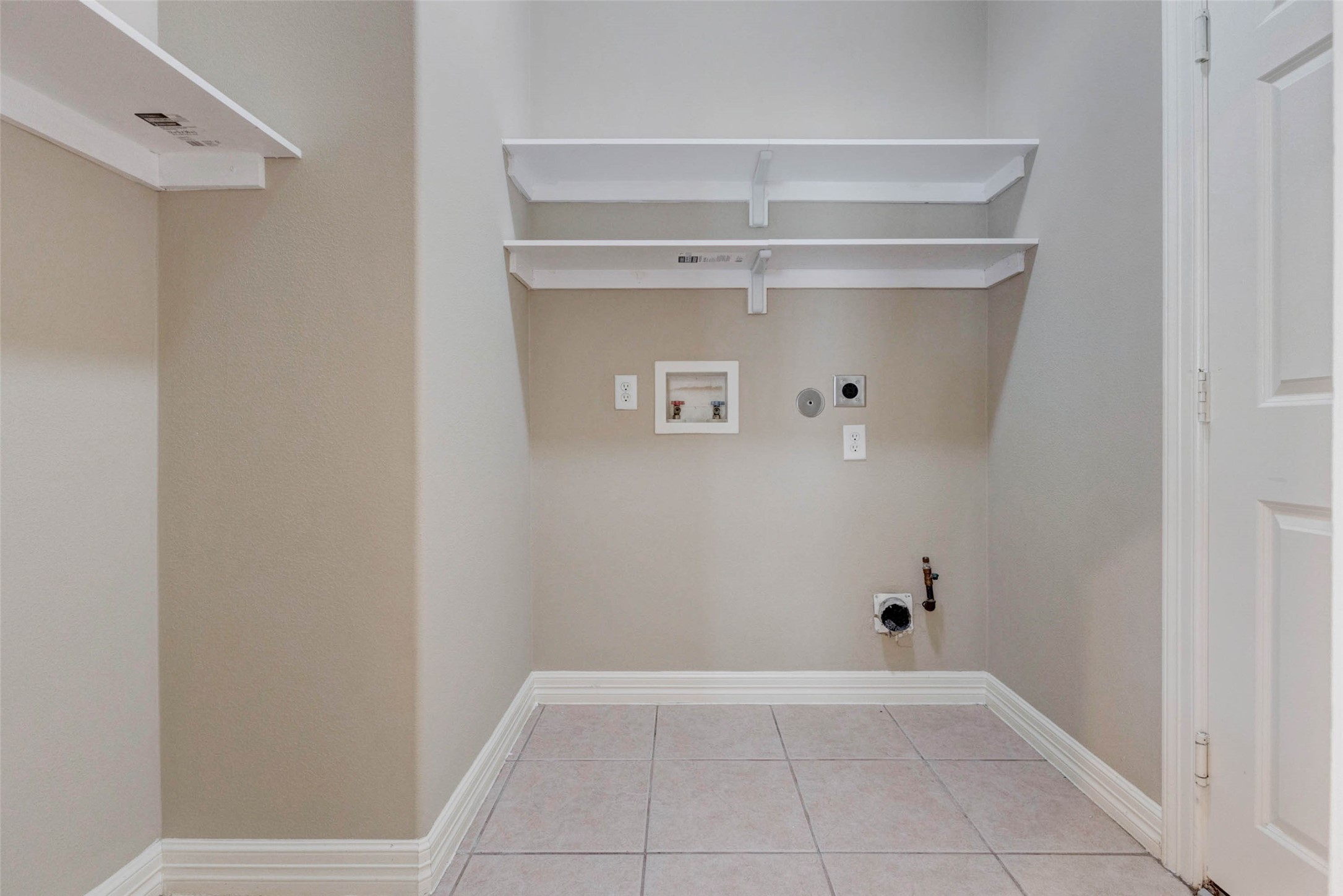 2718 Ridgeglen Lane Houston, TX 77082 - Photo 22 of 26 In-house utility room with tile flooring, washer and dryer connections, built-in shelving and a practical recessed laundry area. The hookups are positioned beneath two open shelves that provide space for detergent, supplies and household essentials. A nearby side shelf adds even more storage potential, while the neutral walls, white trim and easy-care flooring keep the room functional, clean and conveniently located inside the home.