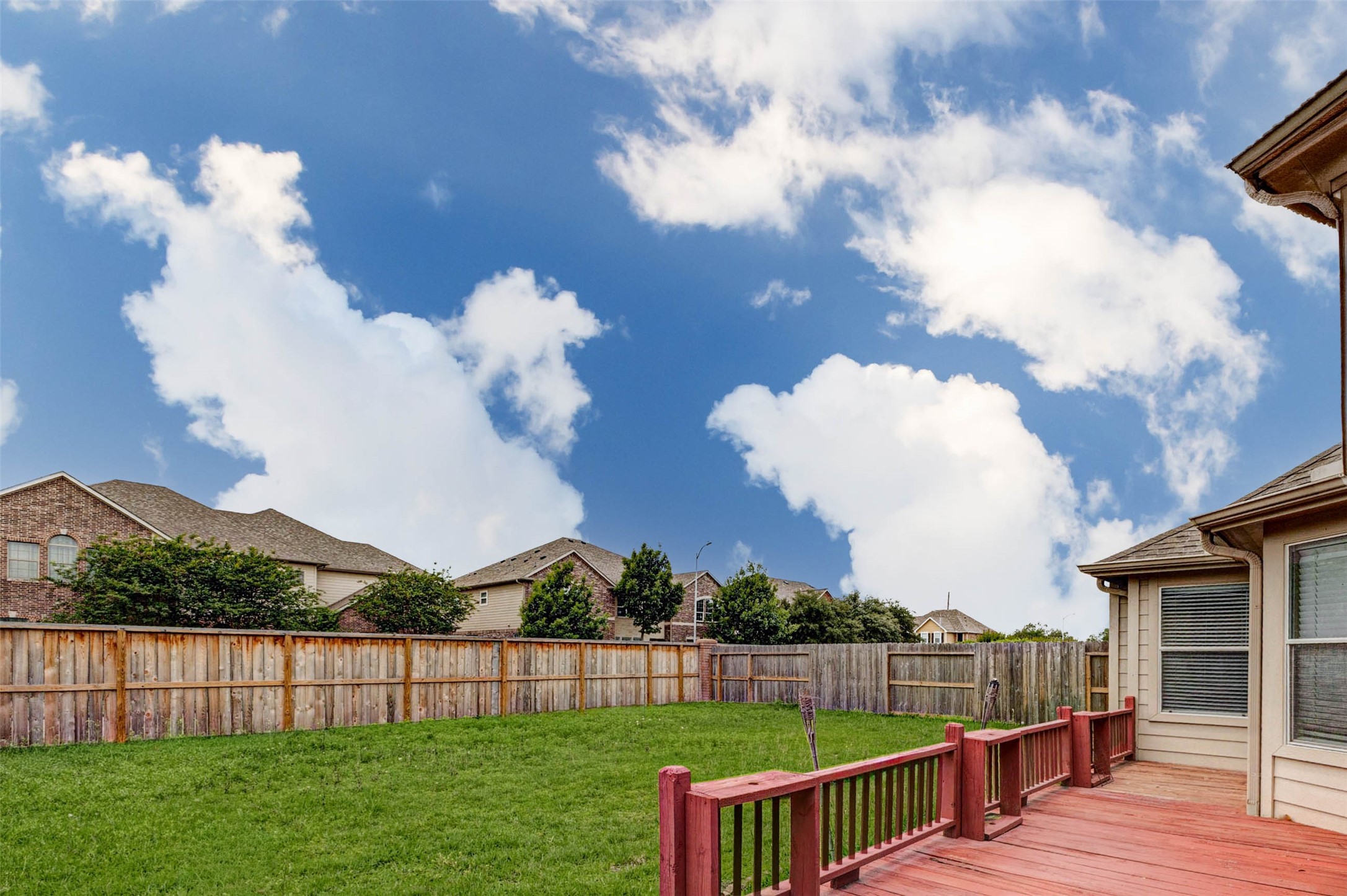 2718 Ridgeglen Lane Houston, TX 77082 - Photo 23 of 26 Backyard view from the red wood deck overlooking a fenced green lawn and wide open sky. The outdoor space offers room for pets, play, container gardening or casual relaxation, with a full wood privacy fence wrapping the yard. The deck creates a natural spot for patio furniture, grilling or enjoying fresh air, while the surrounding homes and bright clouds give the space an open neighborhood feel.
