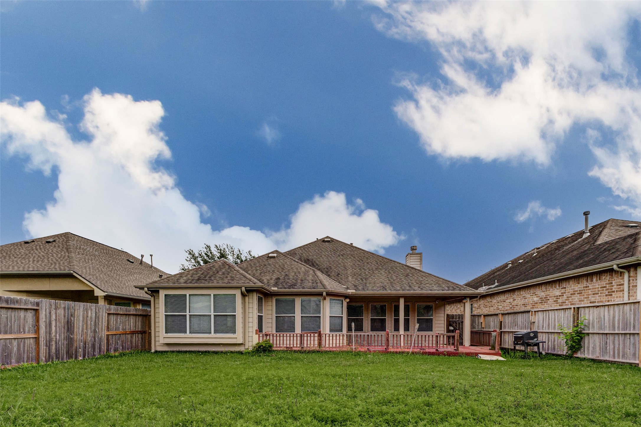 2718 Ridgeglen Lane Houston, TX 77082 - Photo 24 of 26 Rear exterior view showing the single-story home, fenced backyard, green lawn and red wood deck stretching along the back of the house. Multiple rear windows bring natural light into the interior, while the deck offers an easy outdoor extension for seating or entertaining. The fully fenced yard provides usable open space, and the wide view highlights the home’s low-profile design, rear elevation and private outdoor setting.