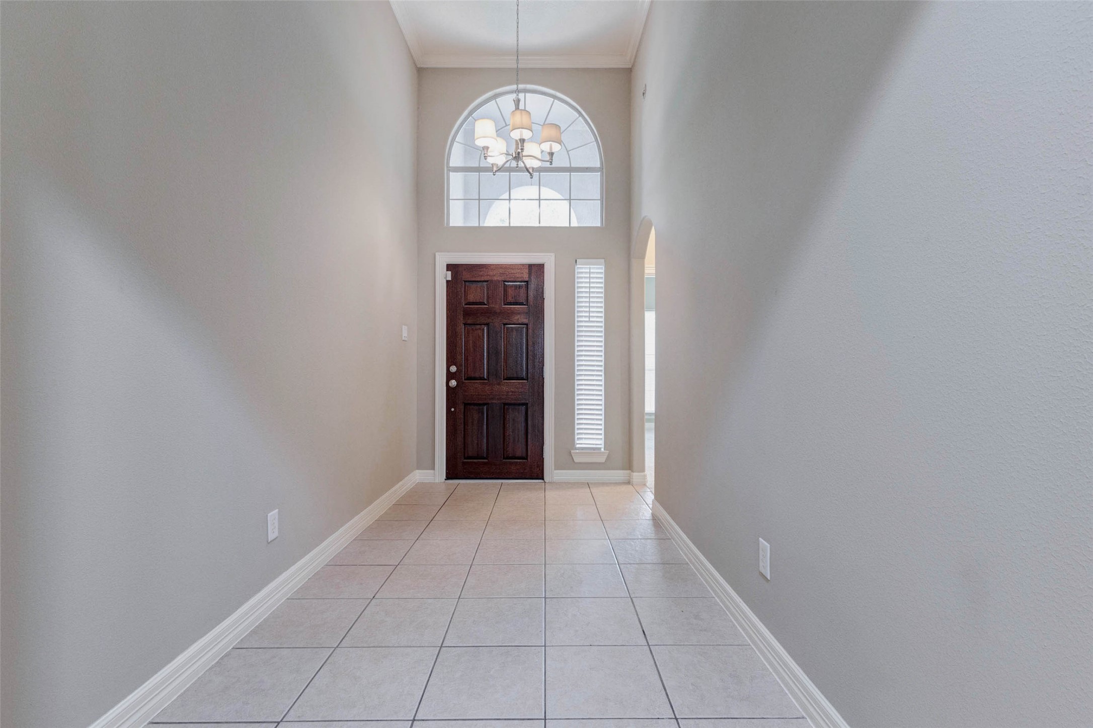 2718 Ridgeglen Lane Houston, TX 77082 - Photo 3 of 26 Bright tiled entry hall with soaring ceiling, crown molding, a chandelier and a dramatic arched transom window above the rich wood front door. A narrow side window with blinds adds natural light while maintaining a polished entry look. The long foyer creates an elegant sense of arrival and leads toward the main living spaces through a graceful arched opening. Neutral walls, white trim and large-format tile establish a clean, refined and memorable first impression.
