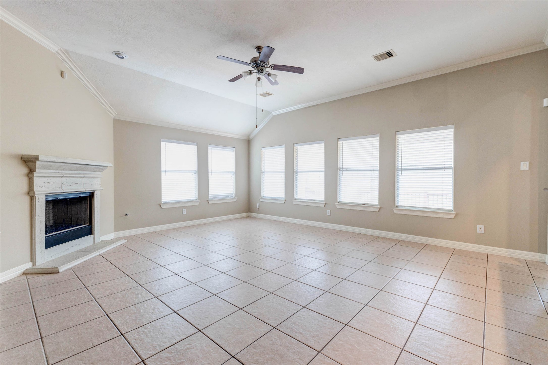 2718 Ridgeglen Lane Houston, TX 77082 - Photo 26 of 26 Spacious main living area with tile flooring, high ceiling, crown molding, ceiling fan and a wall of windows fitted with blinds for filtered natural light. The gas-log fireplace with light stone surround anchors the room, while the open floor plan provides flexibility for seating, dining and everyday living. Neutral walls, bright windows and continuous tile flooring make the space feel open, airy and easy to personalize with a range of furniture styles and décor.