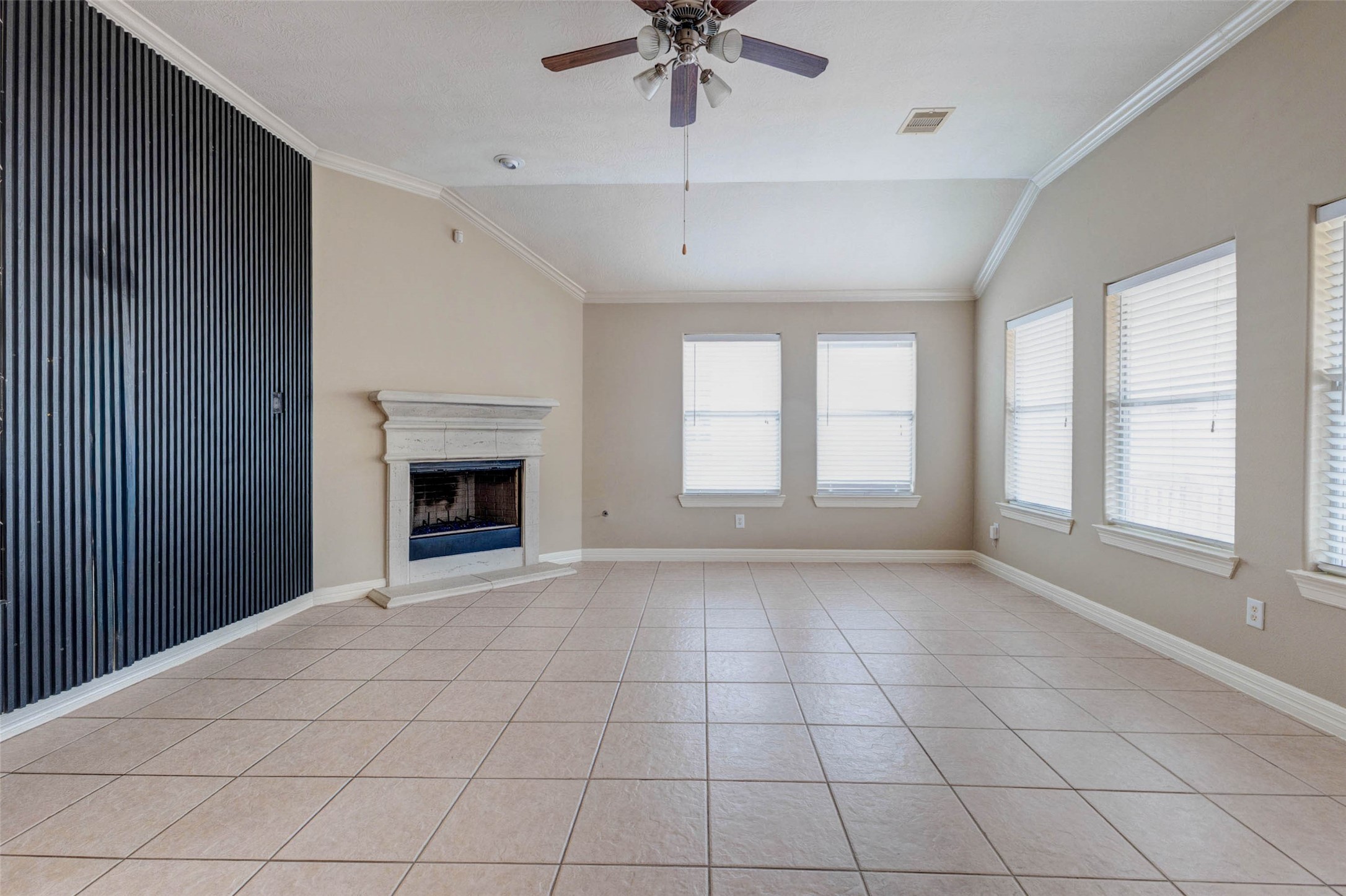 2718 Ridgeglen Lane Houston, TX 77082 - Photo 4 of 26 Living room view highlighting the bold black slatted accent wall and fireplace focal point. The high ceiling, crown molding and ceiling fan add comfort and scale, while multiple windows brighten the room with soft natural light. The textured feature wall creates a dramatic design statement and gives the living area a modern, memorable look against the neutral tile flooring, soft wall tones and classic fireplace surround. A standout space with depth, contrast and personality.