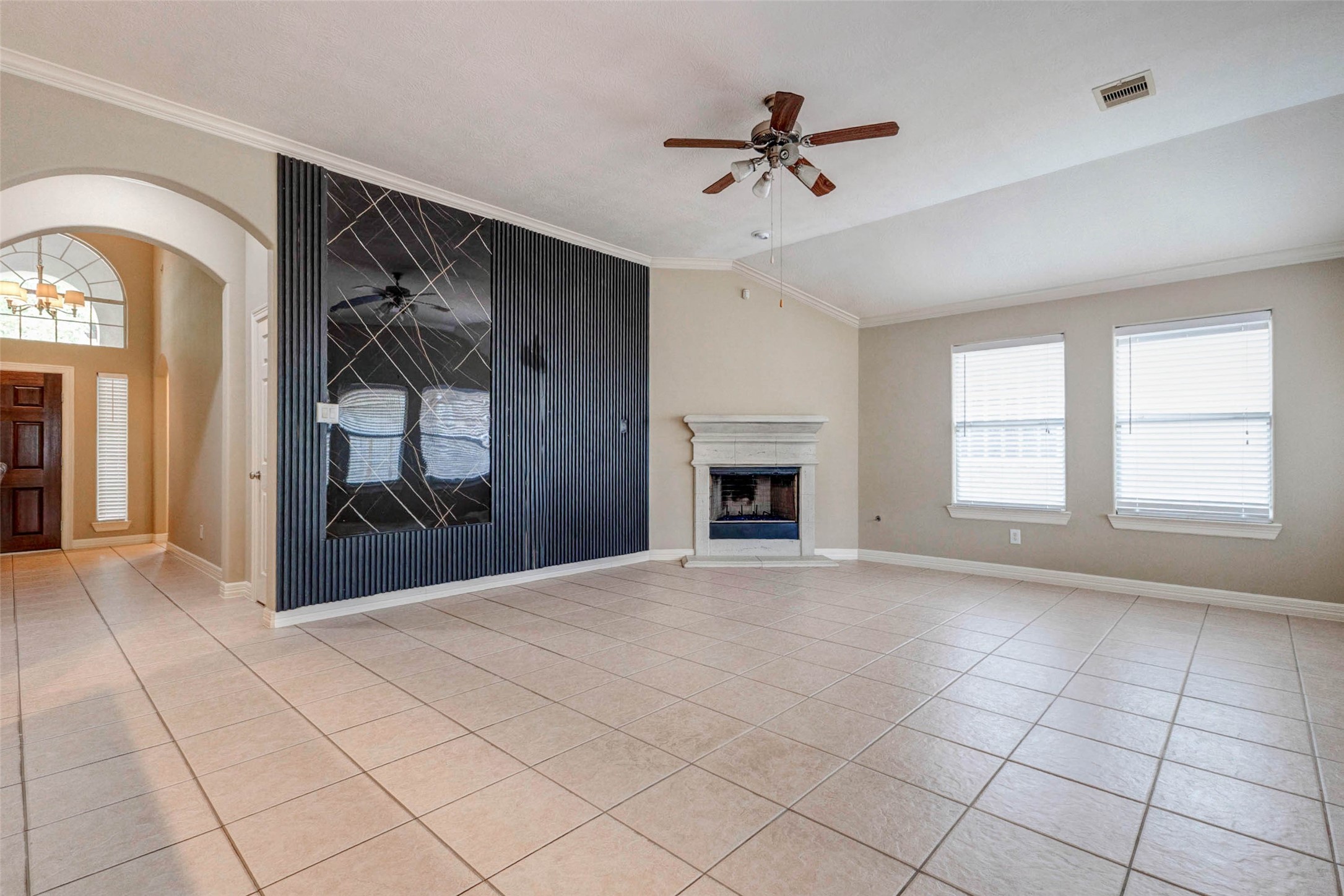 2718 Ridgeglen Lane Houston, TX 77082 - Photo 5 of 26 Open living area with a stylish black accent wall, marble-look inset detail and gas-log fireplace with light stone surround. The arched entryway and front foyer are visible, showing the home’s easy flow from the front door into the main gathering space. Tile flooring, neutral paint, crown molding and high ceilings create a clean, polished setting with room for comfortable seating, media placement and décor. The contrast wall adds depth, definition and visual interest.