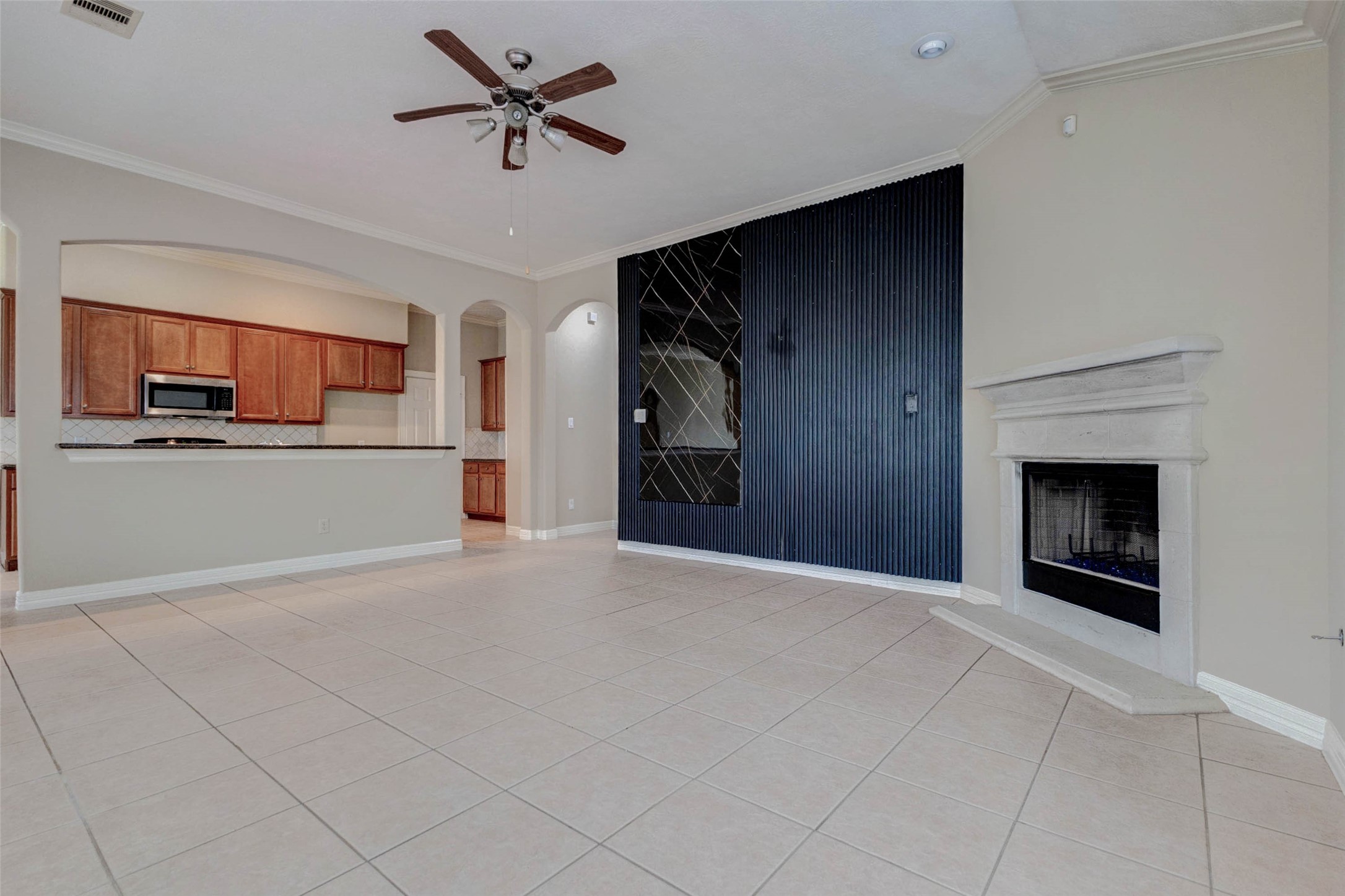 2718 Ridgeglen Lane Houston, TX 77082 - Photo 6 of 26 Wide living room angle showing the open connection to the kitchen through a raised breakfast bar. The fireplace sits to one side, while the dramatic black accent wall adds visual depth and contrast. Arched openings, tile flooring, crown molding and a ceiling fan enhance the room’s architectural interest, making the space feel open, functional and well connected for daily living, casual dining and entertaining. The layout keeps the kitchen in clear view and conversation close.