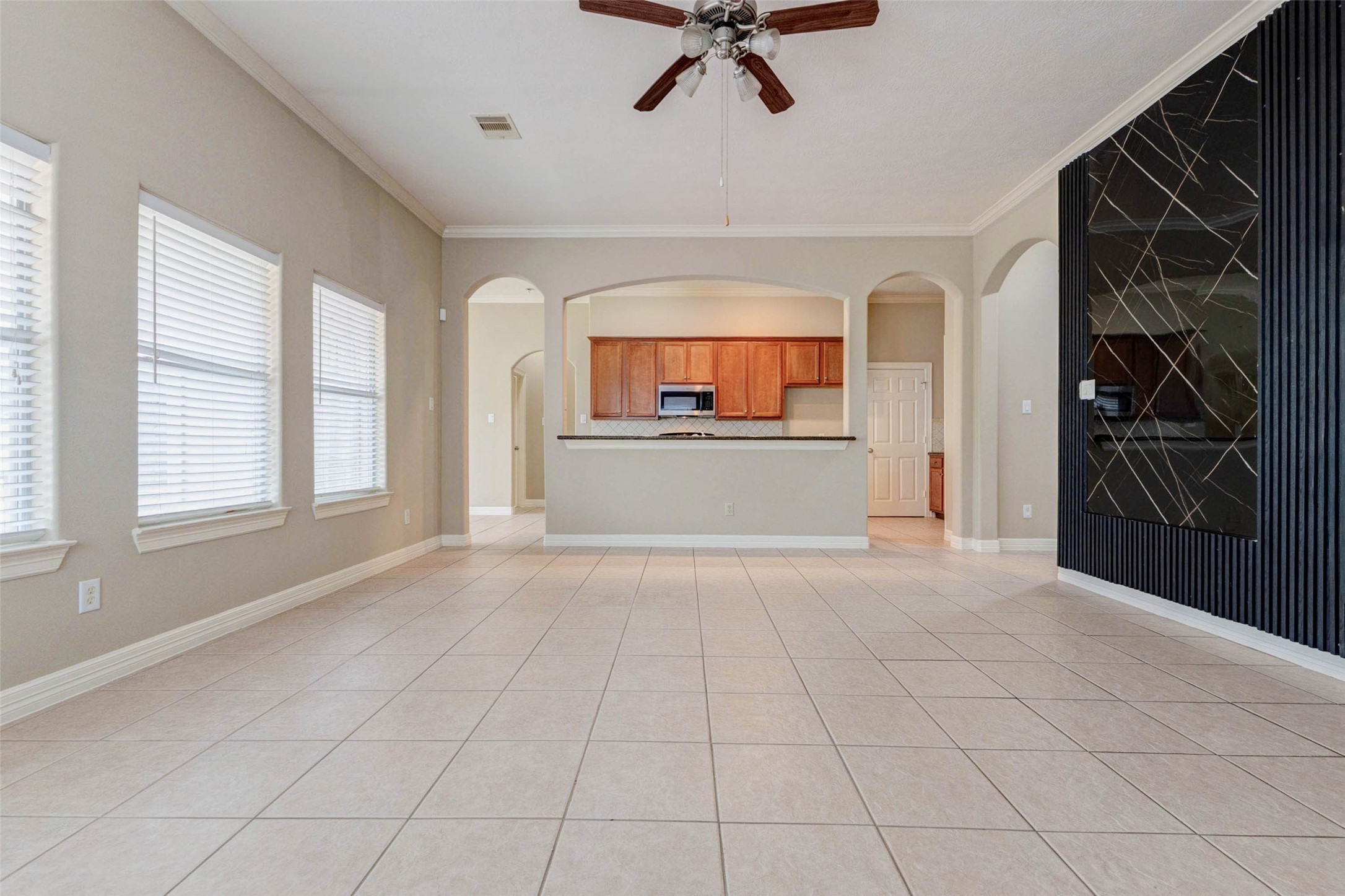 2718 Ridgeglen Lane Houston, TX 77082 - Photo 7 of 26 View across the living and dining area toward the kitchen, emphasizing the home’s open-concept flow and abundant natural light. A row of windows lines the left wall, while the breakfast bar and warm wood kitchen cabinetry sit straight ahead. The black accent wall adds a contemporary focal point, and the tiled floor provides a continuous, low-maintenance surface that visually connects the living, dining and kitchen areas. A flexible space for everyday use and entertaining.