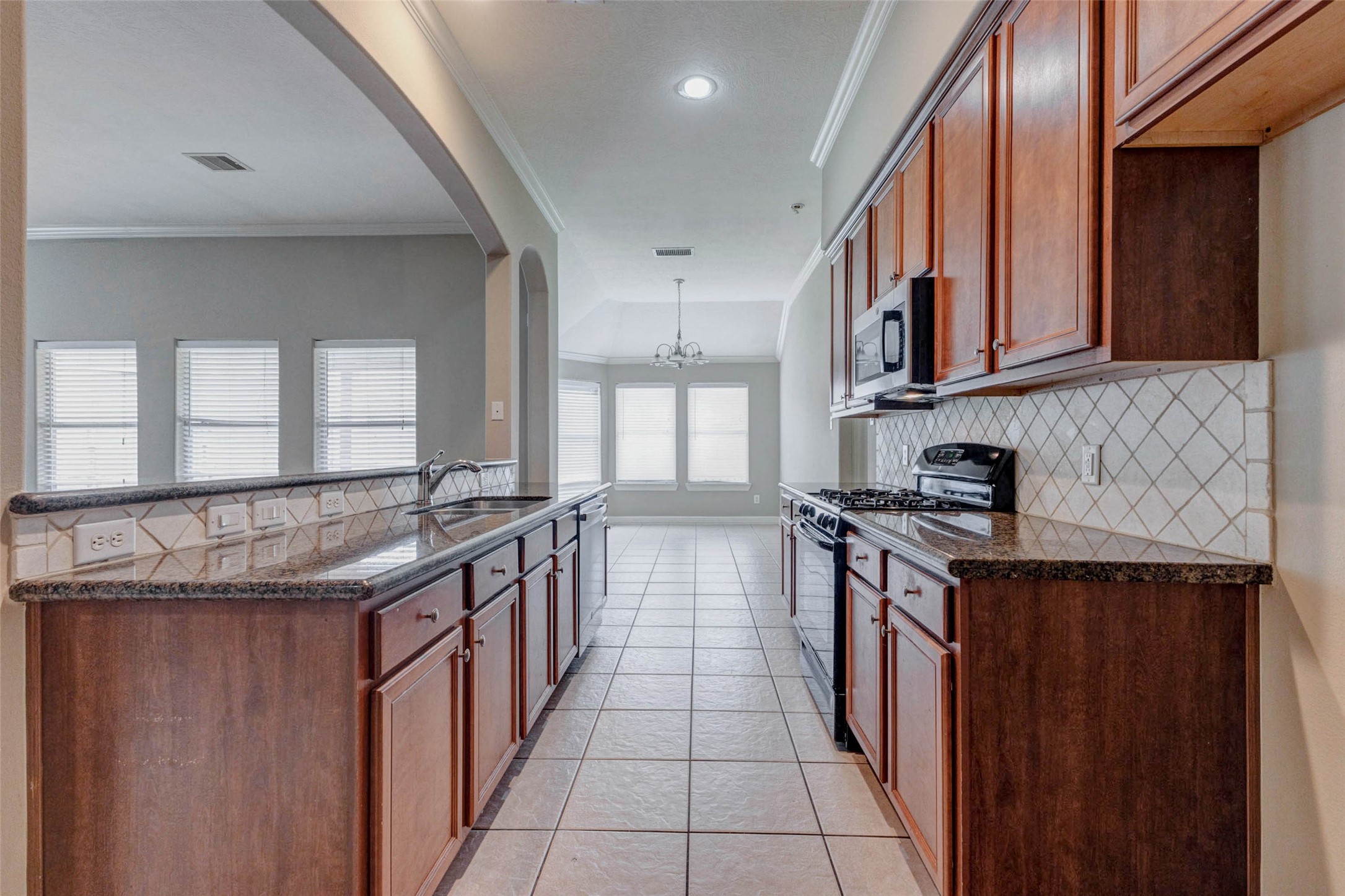 2718 Ridgeglen Lane Houston, TX 77082 - Photo 8 of 26 Functional kitchen with granite countertops, rich wood cabinetry, tile backsplash, gas range, built-in microwave, dishwasher and an open breakfast bar overlooking the living area. The galley-style layout offers efficient workspace on both sides, while recessed lighting and sightlines into the dining area keep the space bright, practical and connected. Warm cabinet tones pair nicely with the neutral tile and granite surfaces for a polished everyday cooking and serving space.