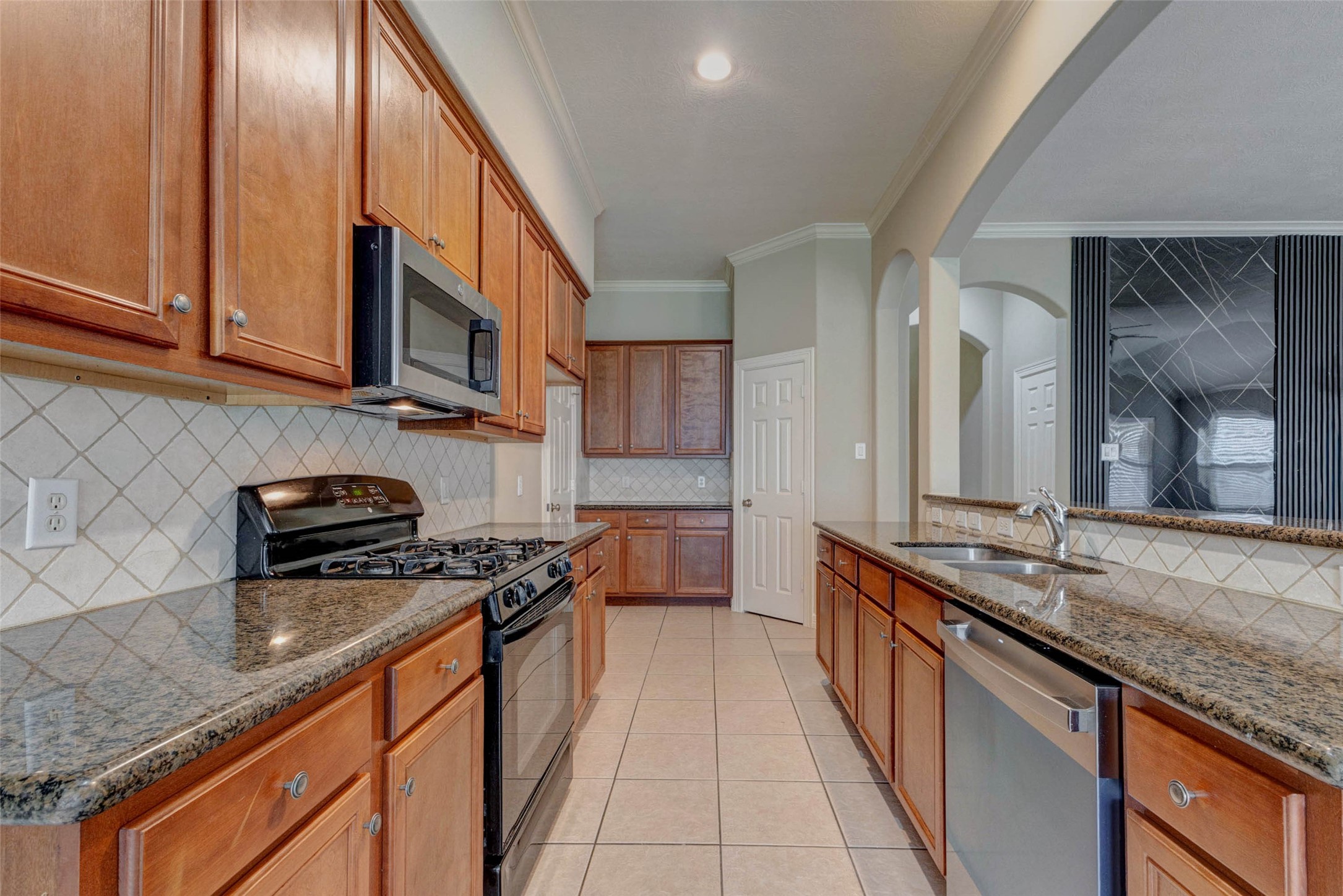 2718 Ridgeglen Lane Houston, TX 77082 - Photo 9 of 26 Well-appointed kitchen with warm wood cabinetry, granite countertops, tile backsplash, gas range, built-in microwave, dishwasher and stainless sink. The galley-style layout offers generous counter space on both sides, while the raised breakfast bar opens to the living area for easy conversation and serving. Arched openings, recessed lighting and a view of the dramatic accent wall create a polished, connected feel.