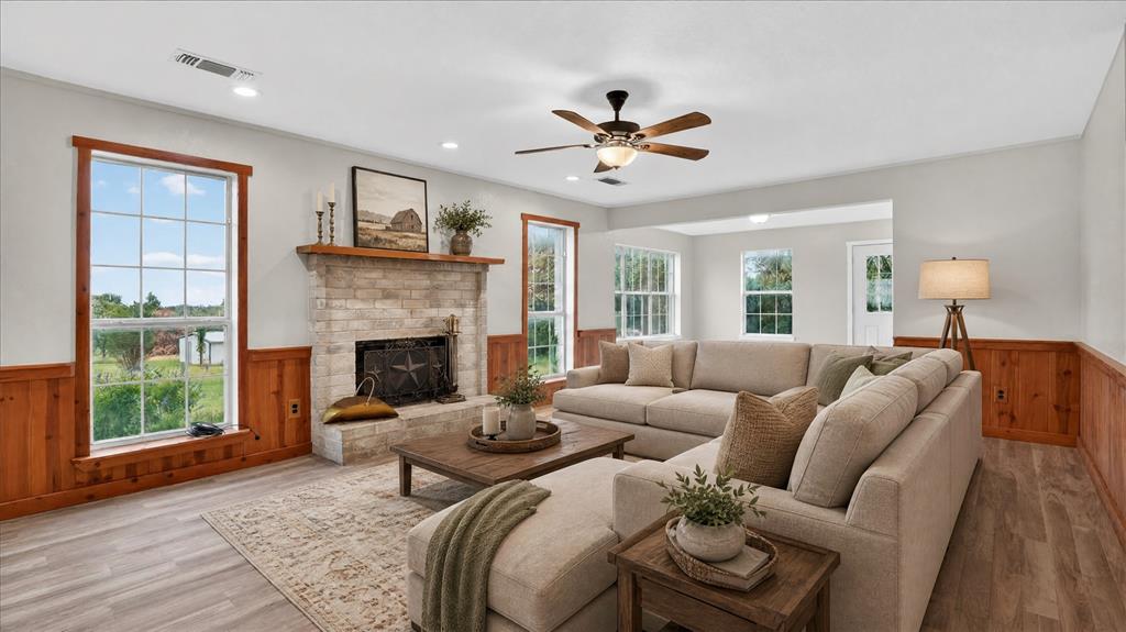 Virtually staged. Spacious living area featuring a stone-clad fireplace with a wood mantle, multiple windows, wood-finish flooring, and wood paneling