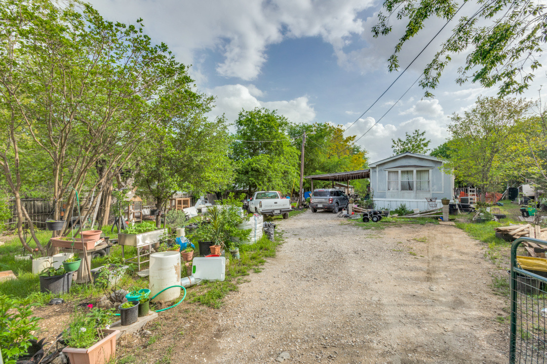 404 Quail Run Buda, TX 78610 - Photo 6 of 13 The property features a gravel driveway, a residential structure with an overhang, and surrounding mature trees with green foliage