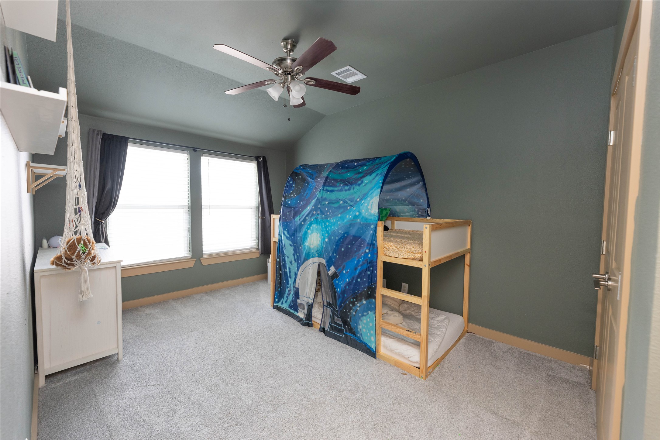 8007 Capitol Street Houston, TX 77012 - Photo 18 of 25 This is a children's bedroom with soft carpeting, featuring a bunk bed with a playful space-themed tent cover. The room is well-lit with a large window and has a ceiling fan for comfort. The walls are painted in a calming gray tone, and there's a small shelf and storage unit for organization.