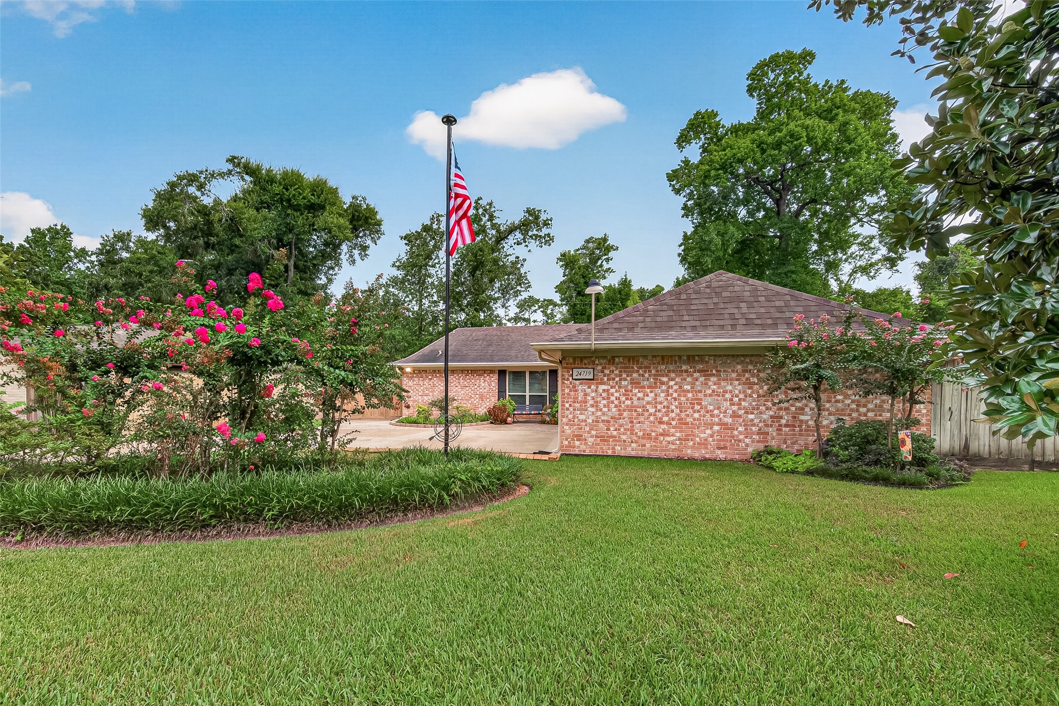 24719 Wilderness Road Spring, TX 77380 - Photo 4 of 42 Charming single-story brick home with a neatly landscaped front yard, featuring vibrant flowering shrubs and a well-maintained lawn. The property includes a driveway leading to the main entrance, and there's a flagpole in the yard. Surrounded by mature trees, it offers a serene and inviting atmosphere.
