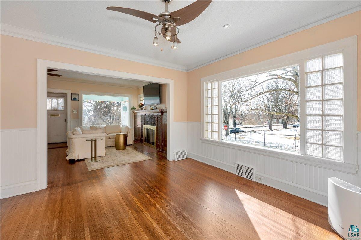 4345 West 8th Street Duluth, MN 55807 - Photo 10 of 31 Unfurnished living room with wainscoting, a ceiling fan, wood-type flooring, ornamental molding, and a glass covered fireplace