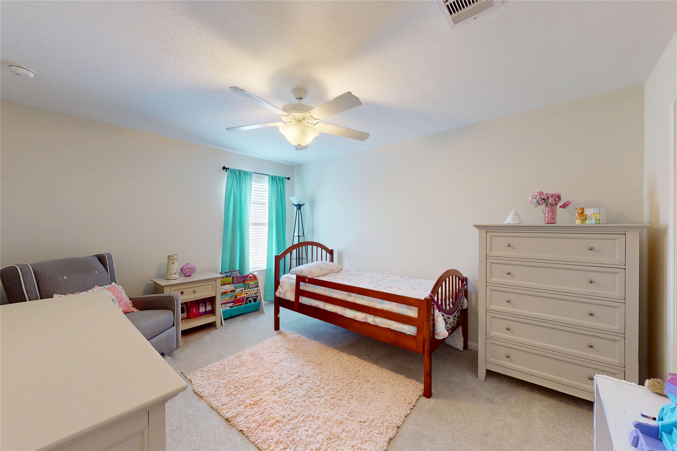 17402 Rainer Valley Lane Humble, TX 77346 - Photo 24 of 33 A cozy, well-lit children's bedroom featuring a single bed, soft carpet, and neutral walls. The room includes a dresser, armchair, and a play area with toys, enhanced by natural light from a window with teal curtains.