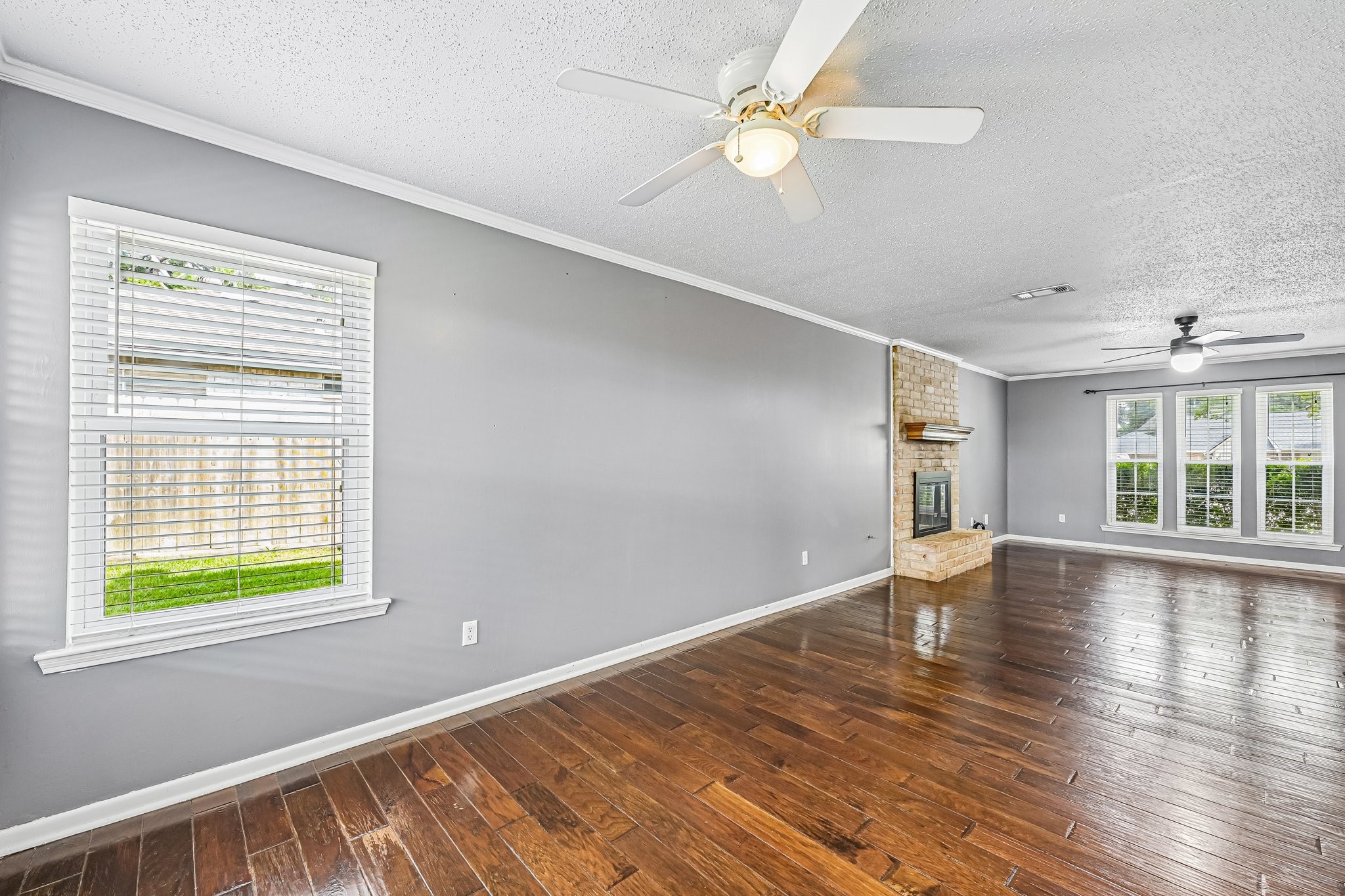 8 Arlington Street Conroe, TX 77301 - Photo 17 of 37 This spacious living room features rich wooden flooring, a cozy brick fireplace, and ample natural light from large windows. The neutral gray walls and ceiling fans add a modern touch.